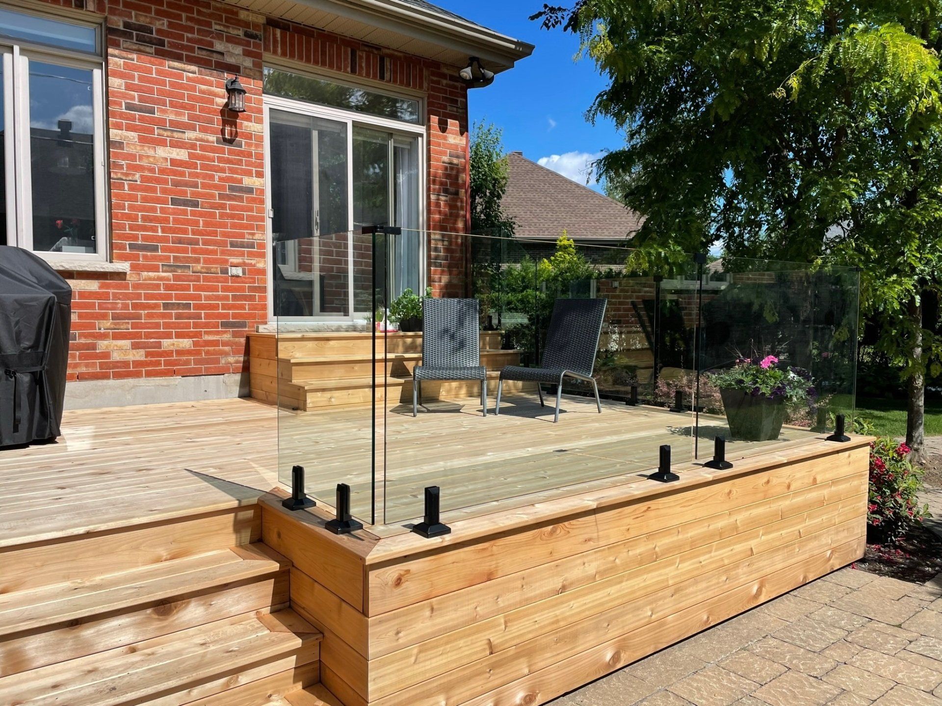 Une terrasse en bois avec une rambarde en verre et des poteaux en métal noir, comprenant un escalier et deux chaises, à l'extérieur d'une maison en briques.