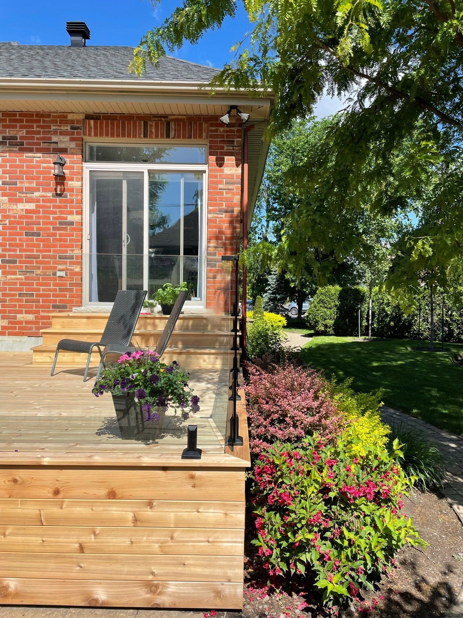 Une terrasse en bois attenante à une maison en briques comprend deux chaises longues, une plante en pot et un escalier menant à un jardin luxuriant.