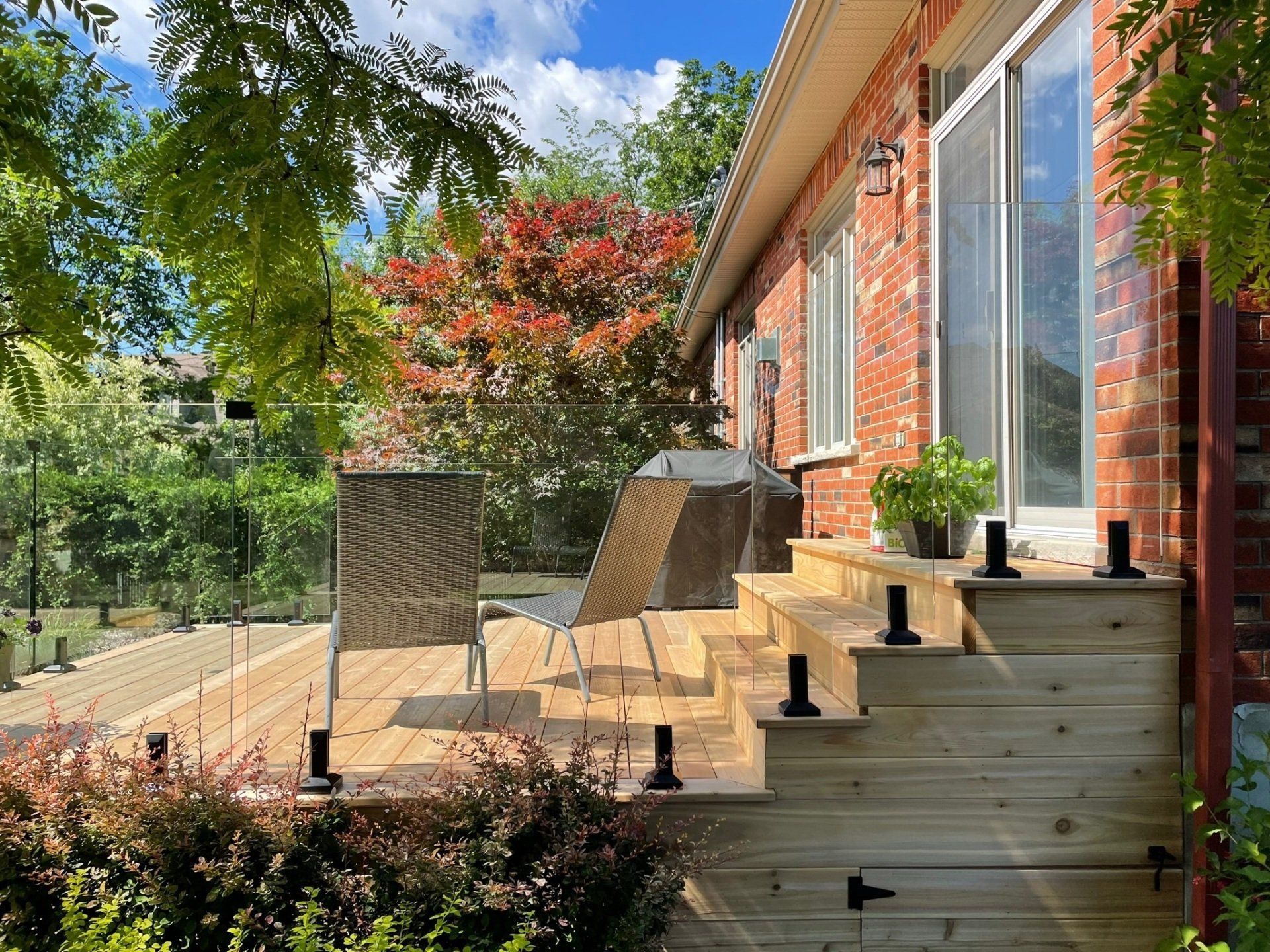 Terrasse en bois avec escalier, deux chaises d'extérieur et une balustrade en verre adossée à un mur de maison en briques rouges.