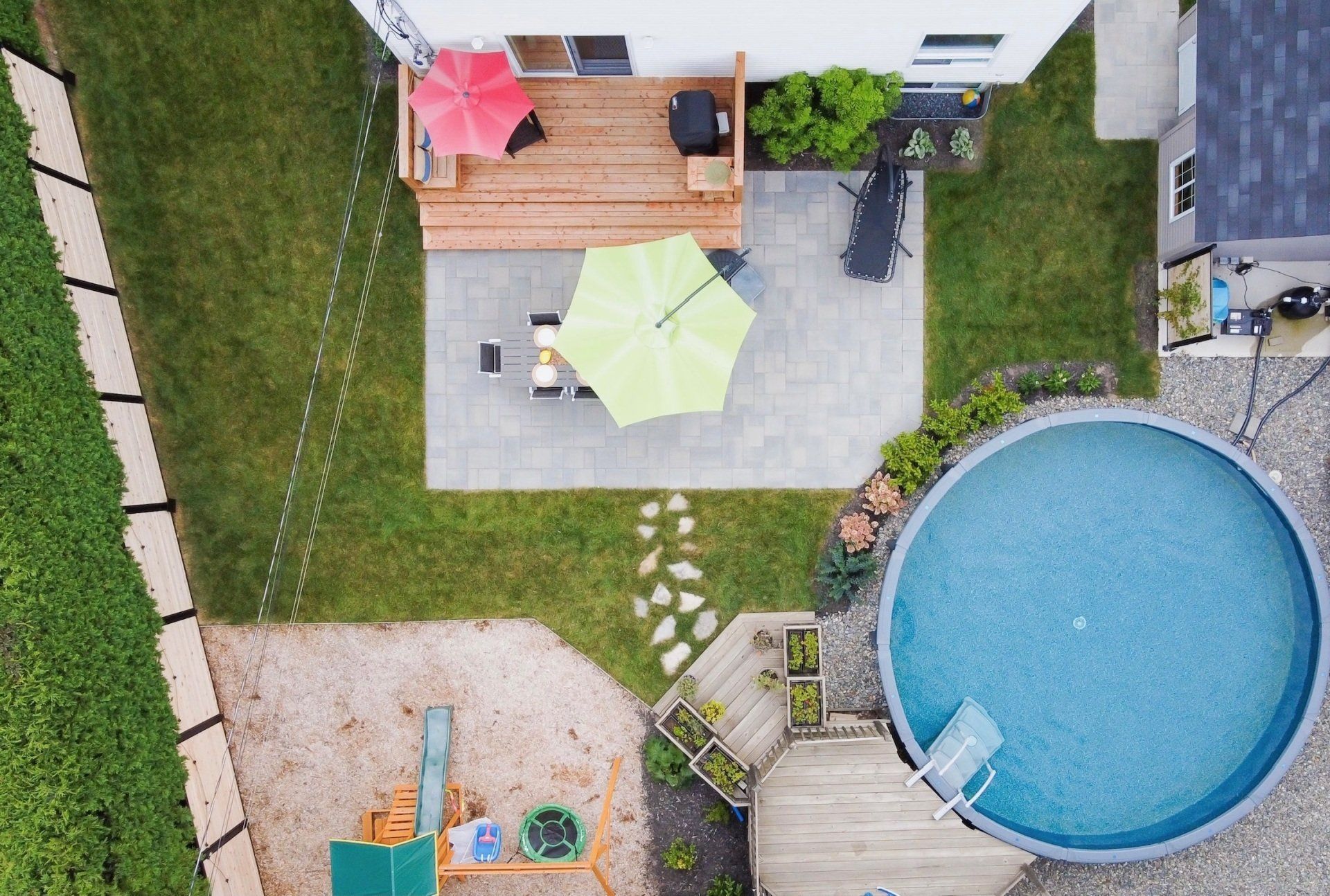 Vue aérienne d'un jardin comprenant une terrasse avec deux parasols, une piscine hors sol ronde et une petite aire de jeux.