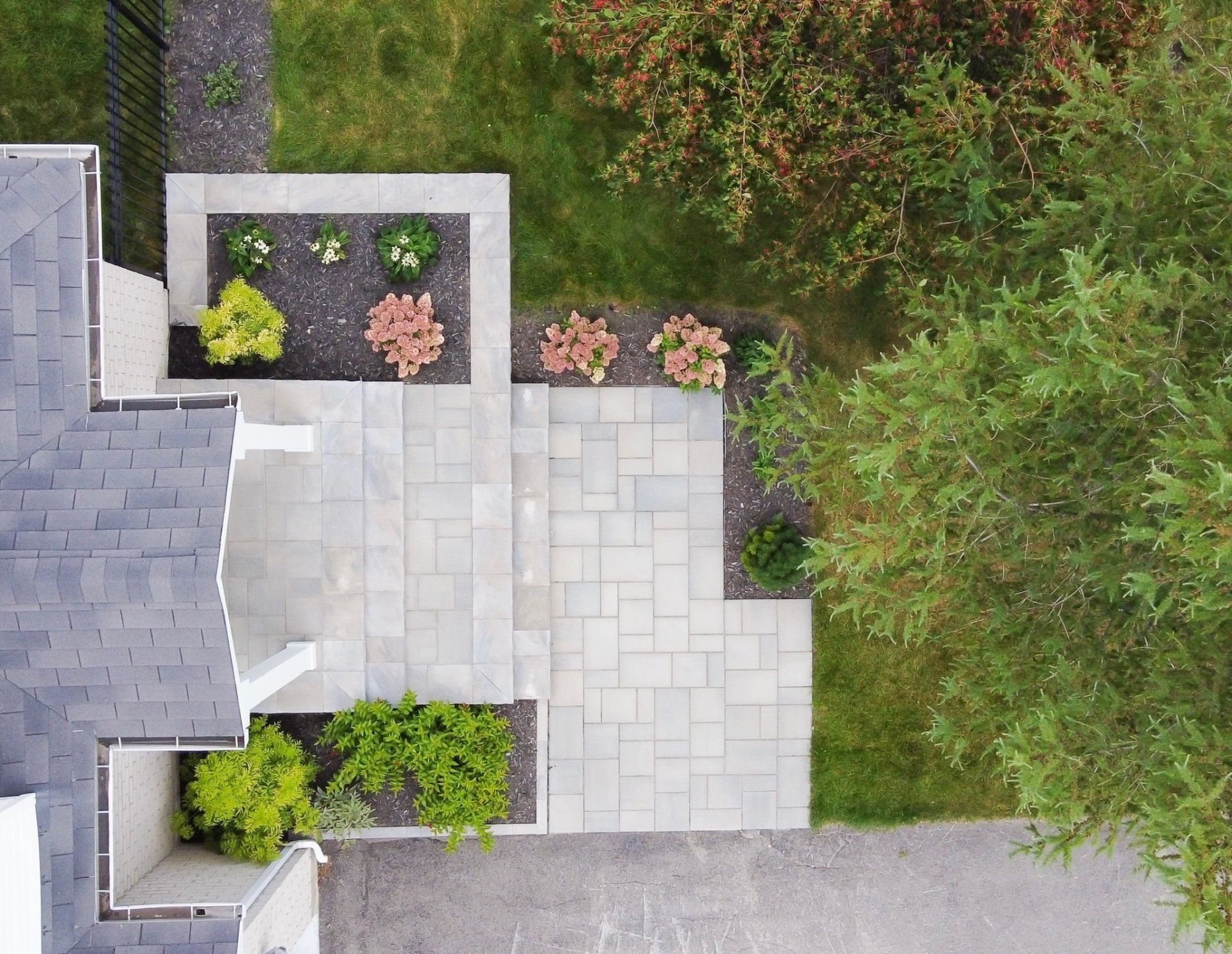 Vue aérienne d'une terrasse en pavés clairs jouxtant une maison avec des parterres de fleurs, des arbustes et une pelouse à proximité.