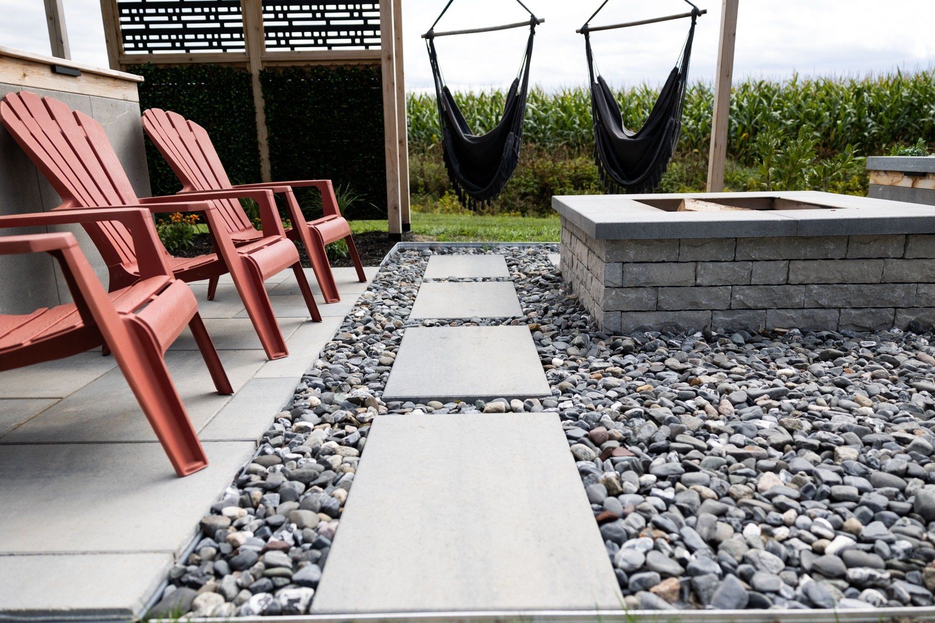Des chaises Adirondack rouges, un foyer en pierre et des chaises suspendues sur une terrasse pavée et recouverte de gravier gris décoratif.