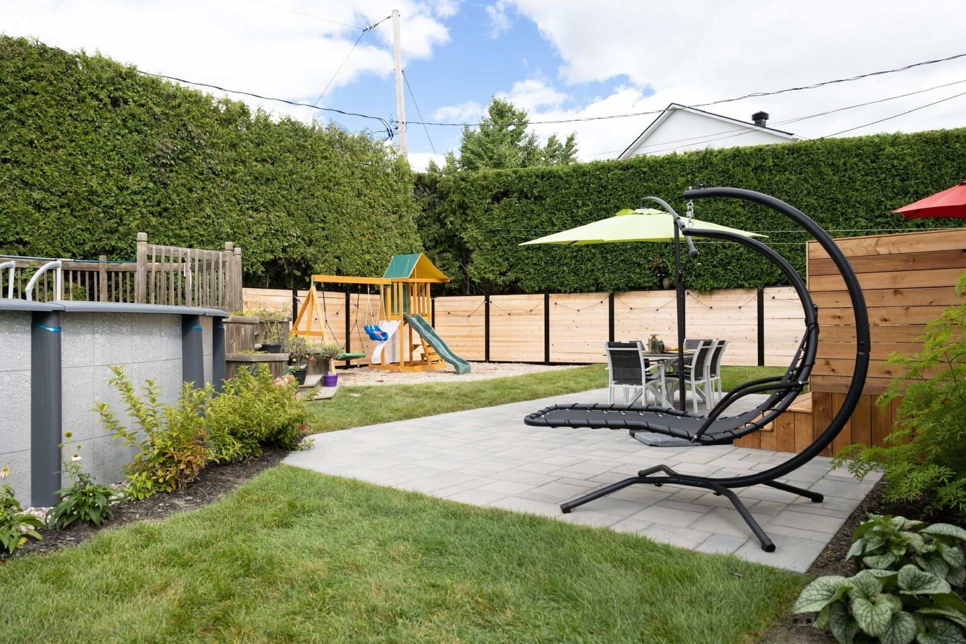 Un jardin avec une terrasse en pierre, un fauteuil suspendu moderne avec parasol, une piscine hors sol et une aire de jeux en bois.