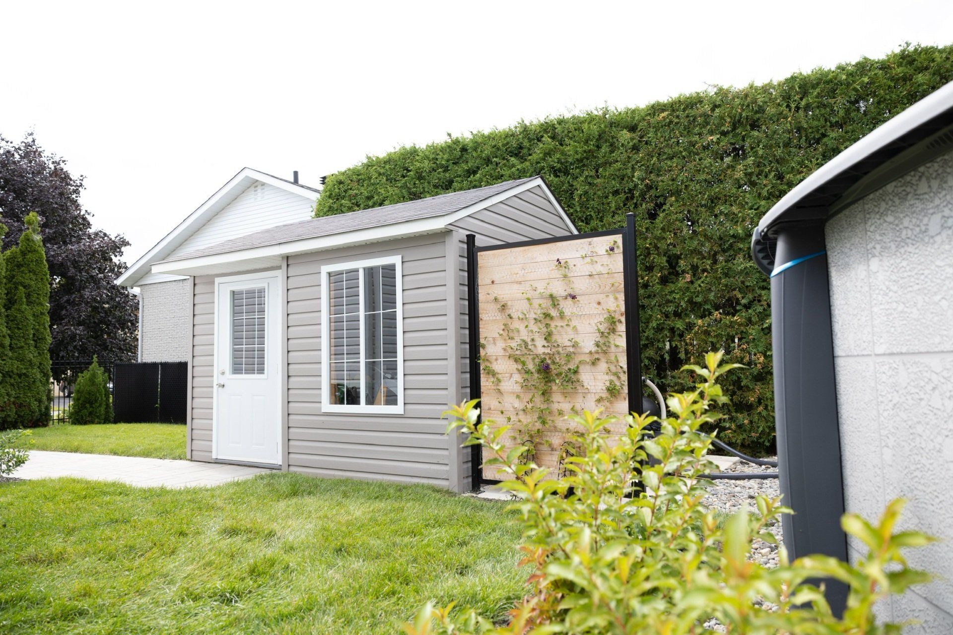Un petit cabanon gris, avec une porte et une fenêtre blanches, se dresse sur une pelouse verte, à côté d'un paravent en bois et d'une haute haie.