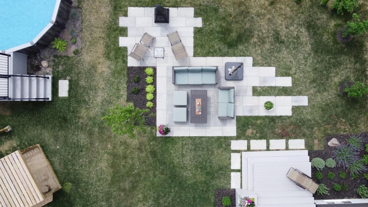 Vue aérienne d'une cour arrière avec une terrasse en pierre, un coin salon extérieur, un foyer et des marches de piscine aménagées dans un jardin gazonné.