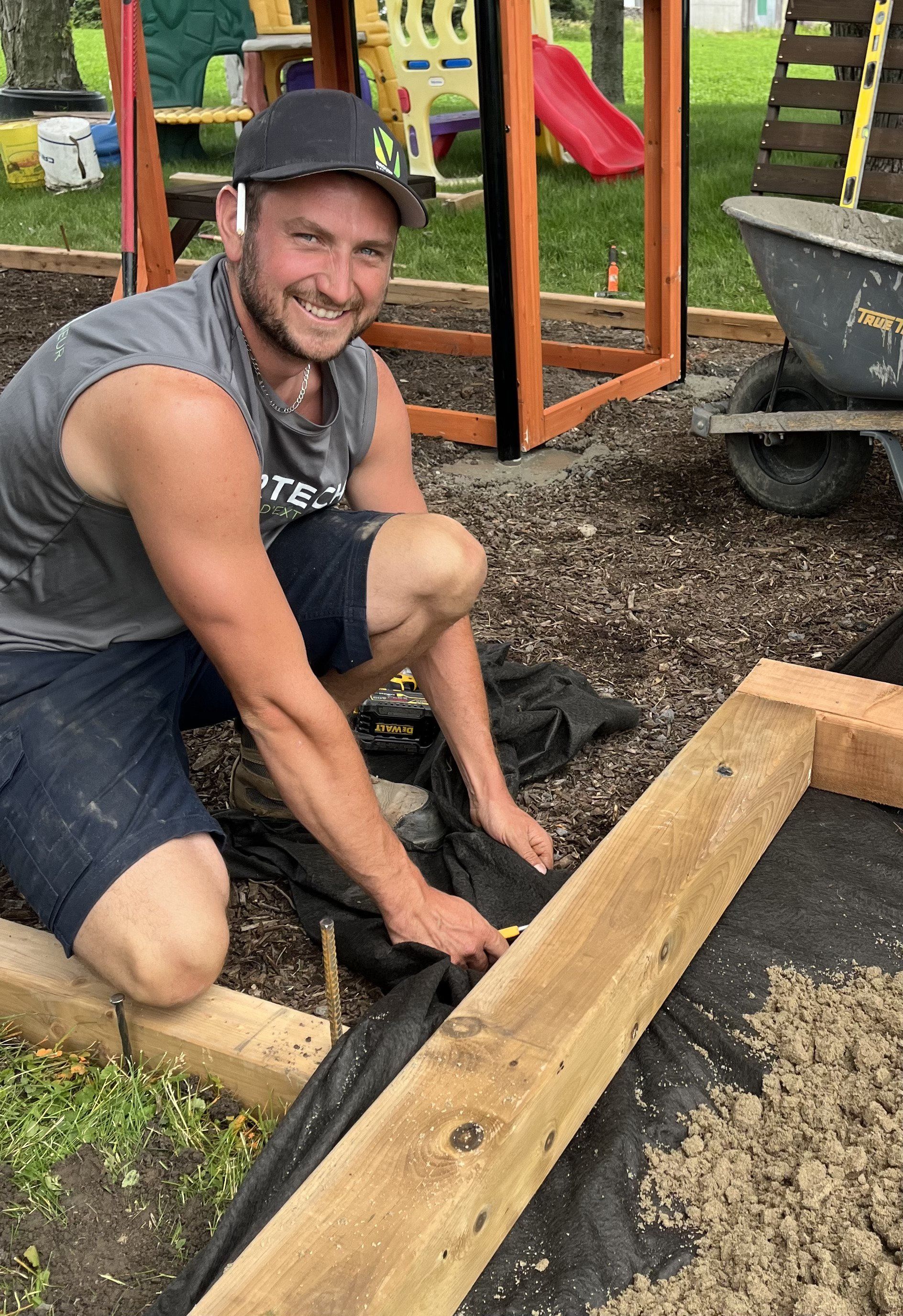 Une personne souriante, vêtue d'un débardeur gris et d'un chapeau, s'accroupit pour installer une toile de paillage contre une ossature en bois.