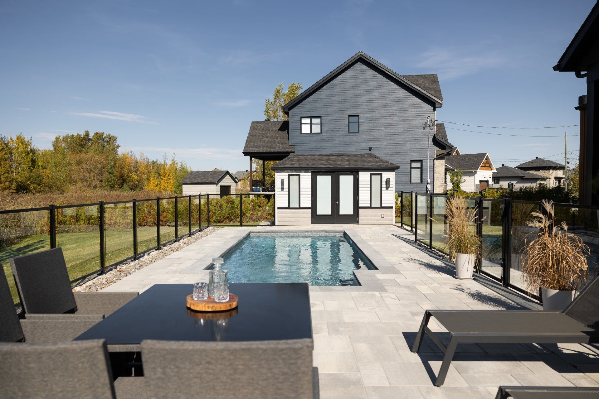 Une terrasse de piscine moderne dans un jardin avec un ensemble de salle à manger noir, une chaise longue, une clôture en verre et un pool house derrière la piscine.
