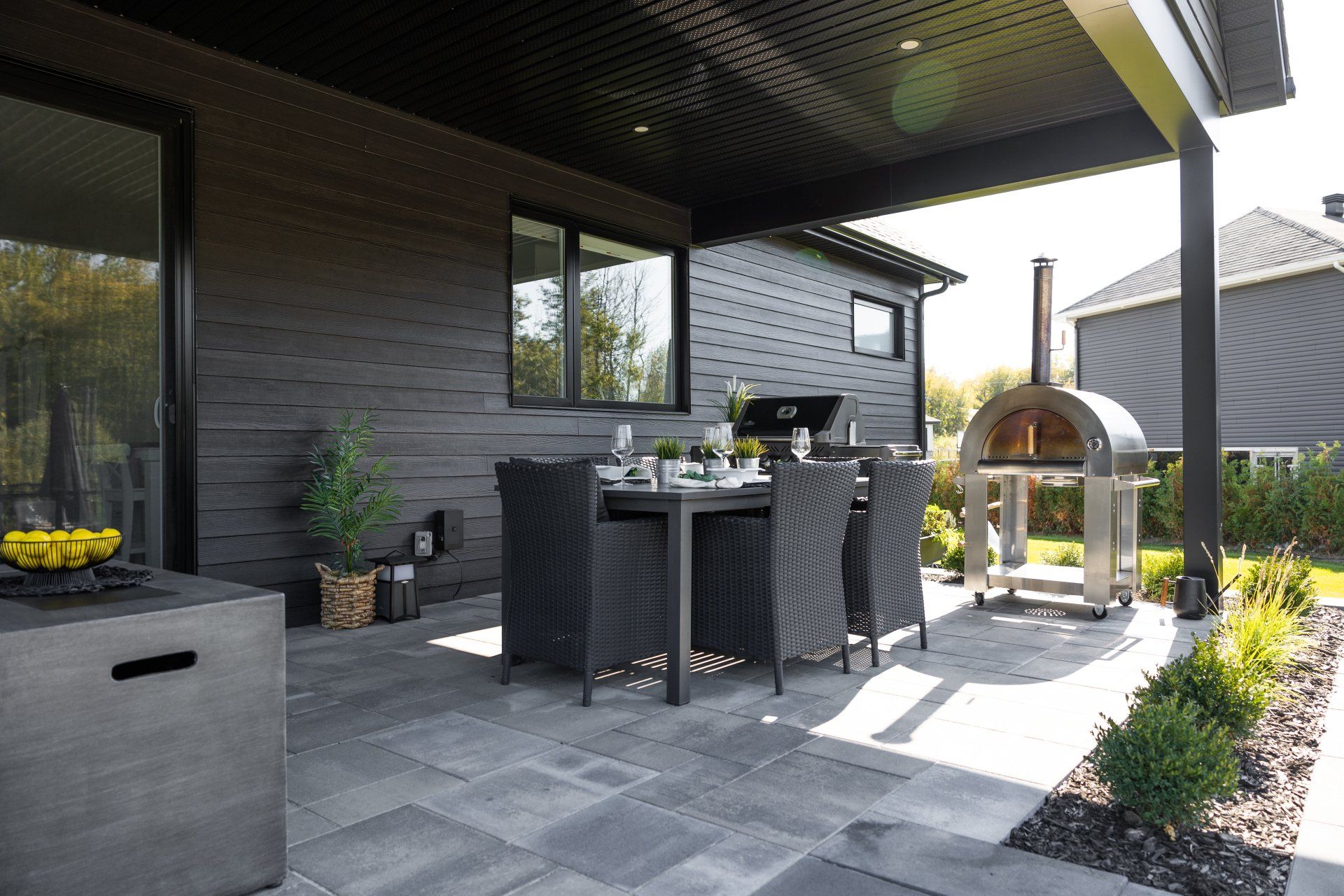 Une terrasse avec chaises longues et un foyer sous une terrasse en bois surélevée, attenante à une maison moderne en pierre et en bois.