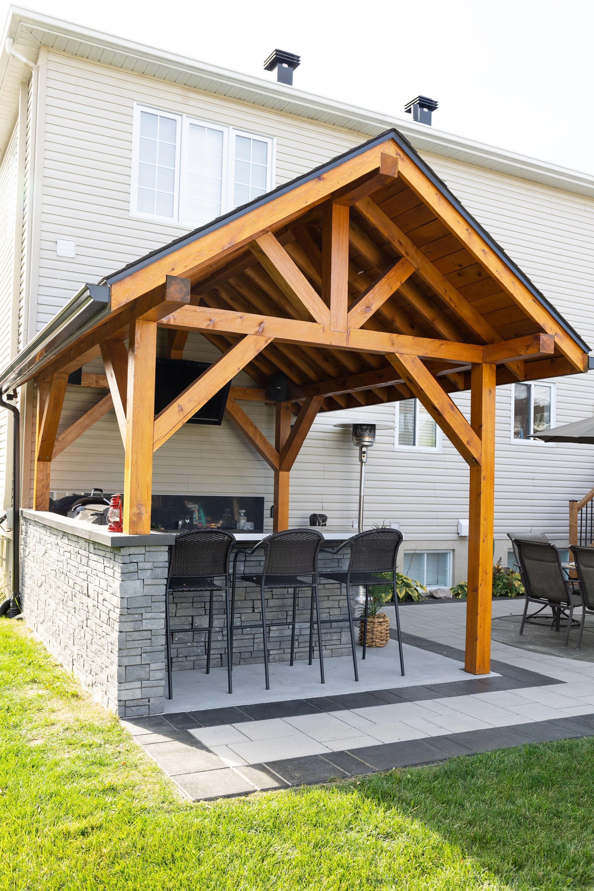 Un pavillon extérieur en bois, avec un bar en pierre et trois chaises hautes, se trouve sur une terrasse pavée attenante à la maison.