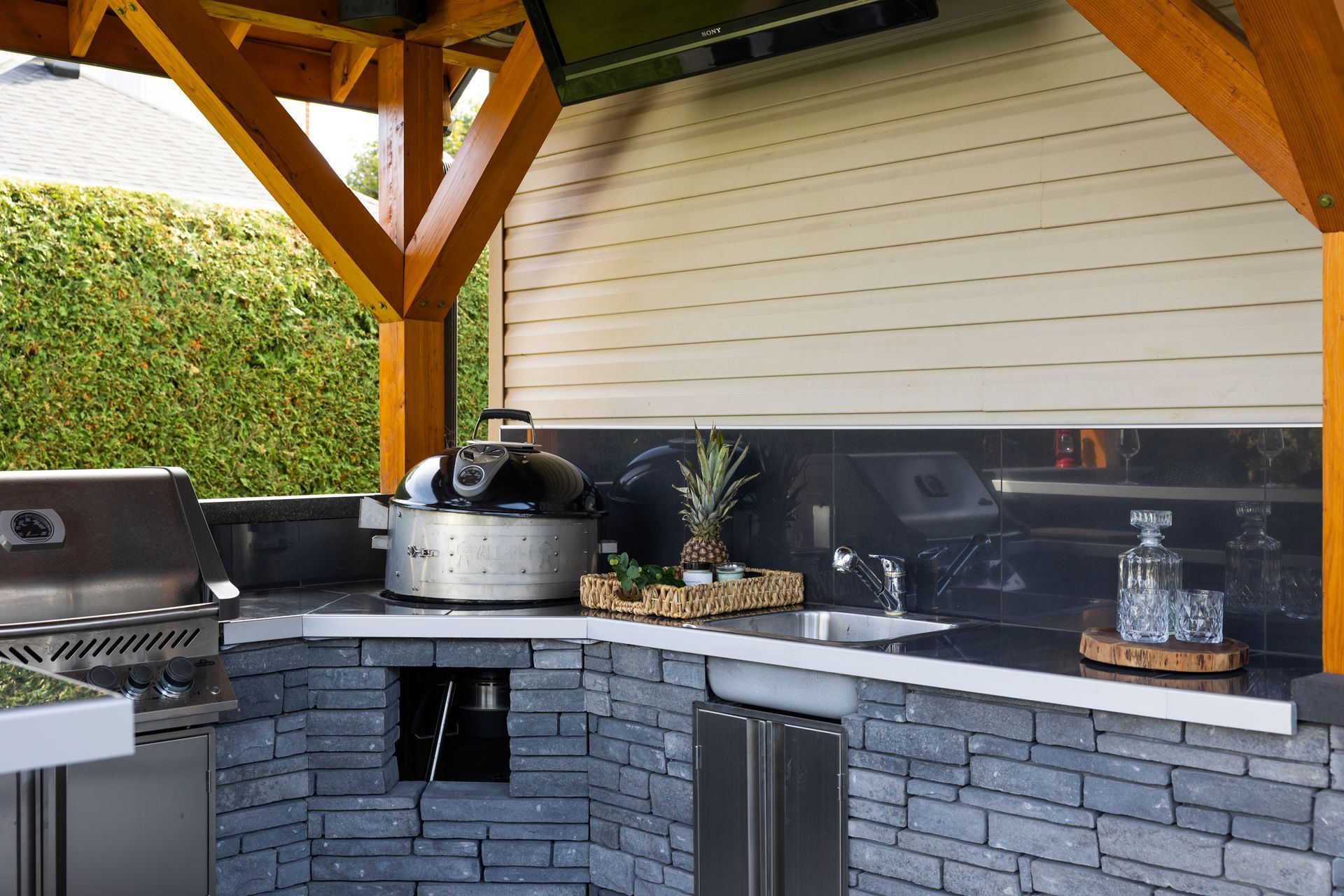 Une cuisine extérieure avec un socle en pierre, un barbecue en acier inoxydable, un évier et une crédence foncée, sous une pergola en bois.