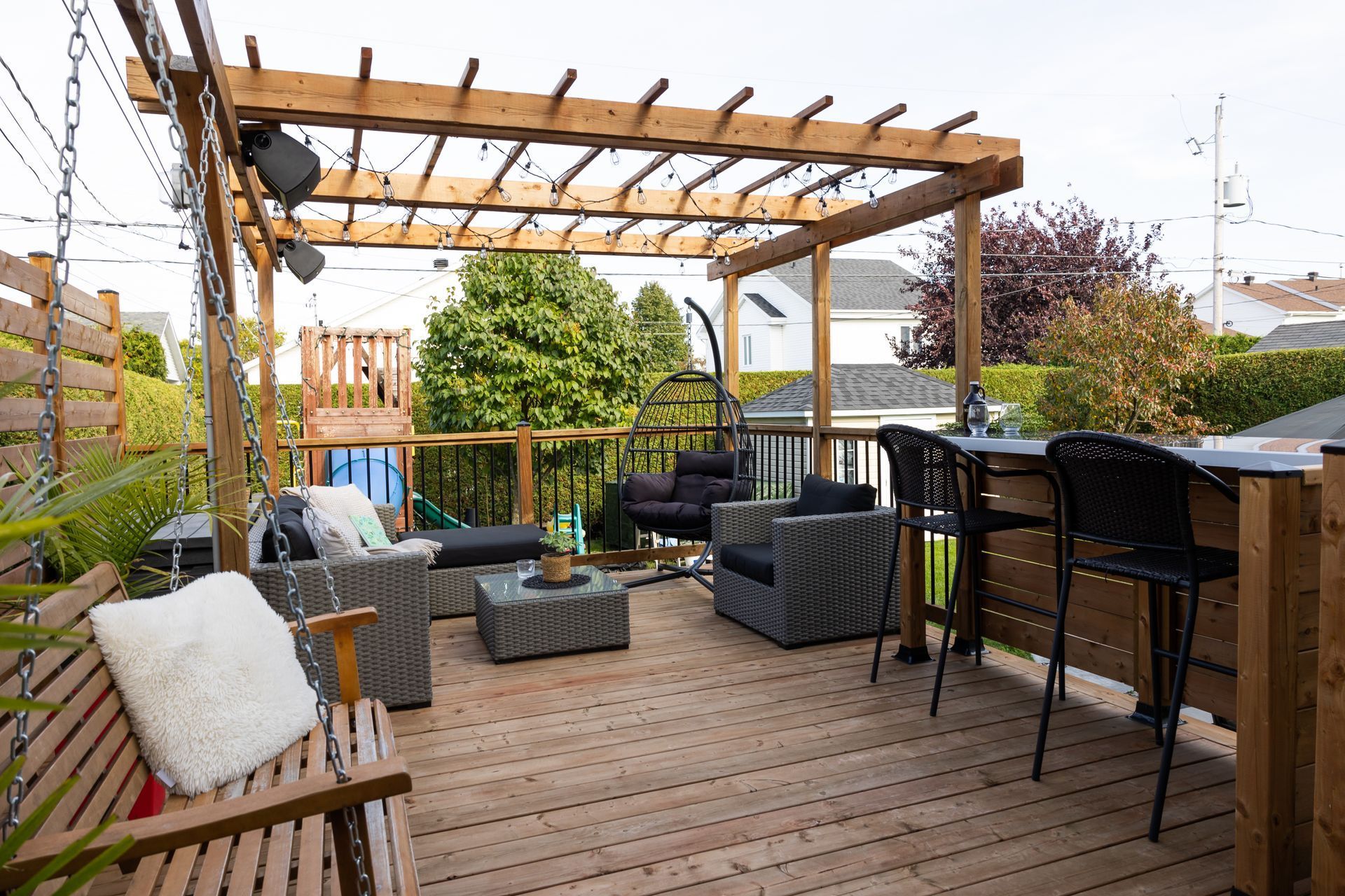 Une terrasse arrière avec une pergola en bois, des chaises longues d'extérieur, une balançoire suspendue et un bar haut avec tabourets.