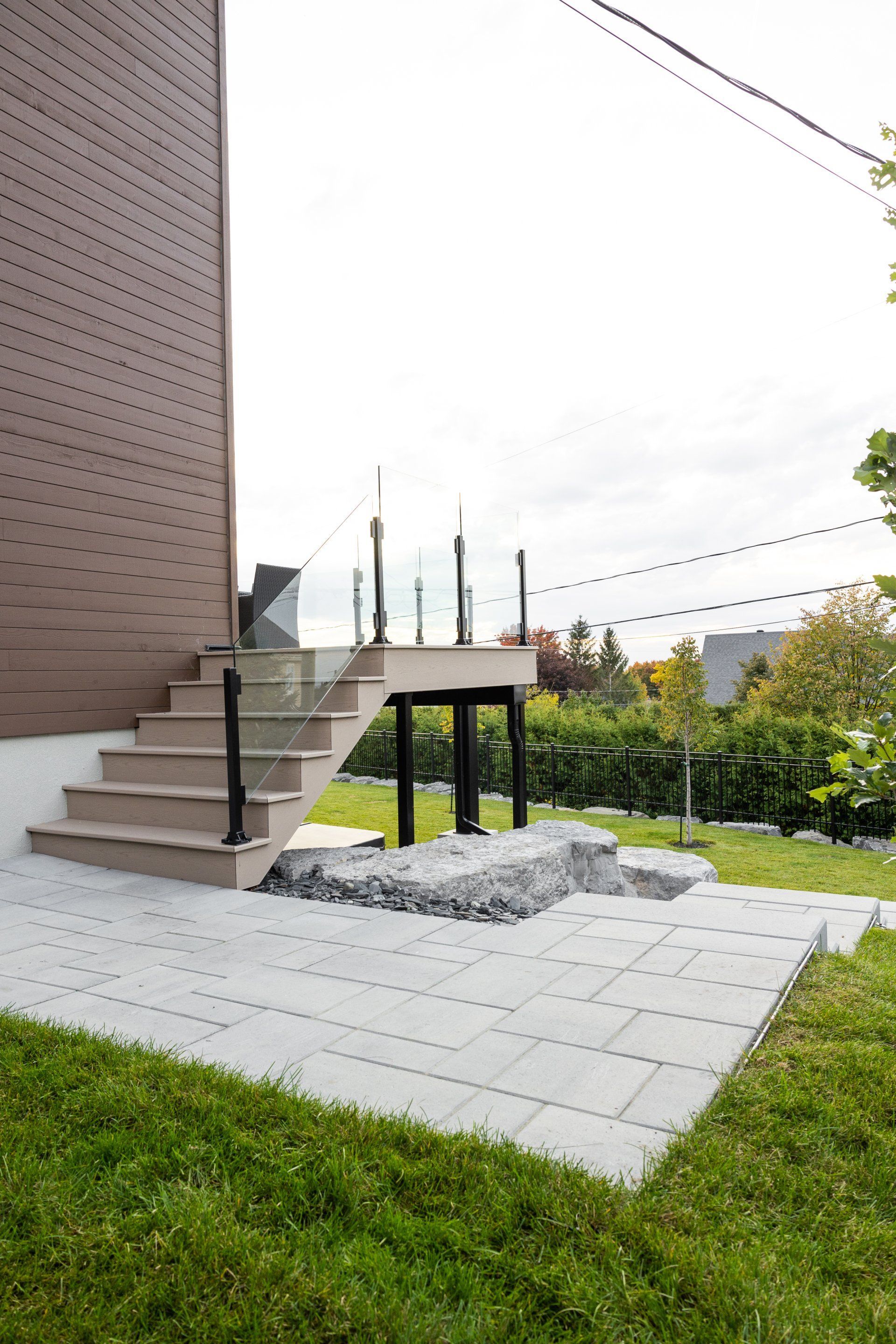 Un patio avec un escalier clair menant à une terrasse surélevée jouxtant une maison brune entourée d'une pelouse verte.
