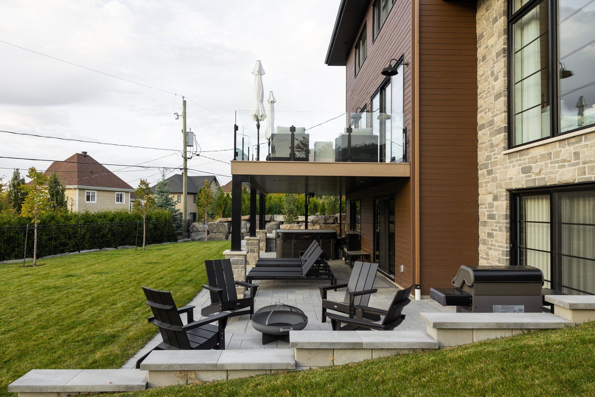 Une maison en pierre avec un patio à l'arrière comprenant des chaises d'extérieur sombres, un foyer et une terrasse surélevée avec balustrade en verre.