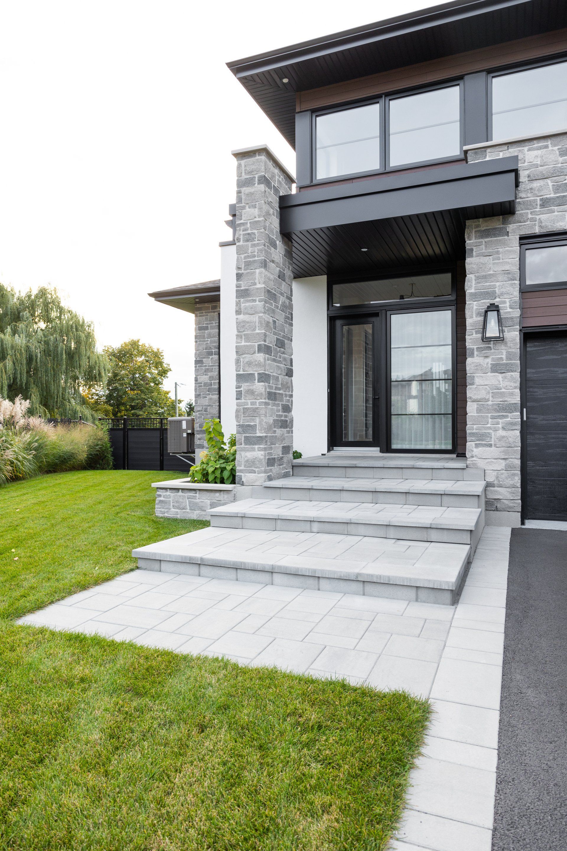 Extérieur de maison moderne avec façade en pierre, entrée à plusieurs marches pavée et pelouse verte bien entretenue.