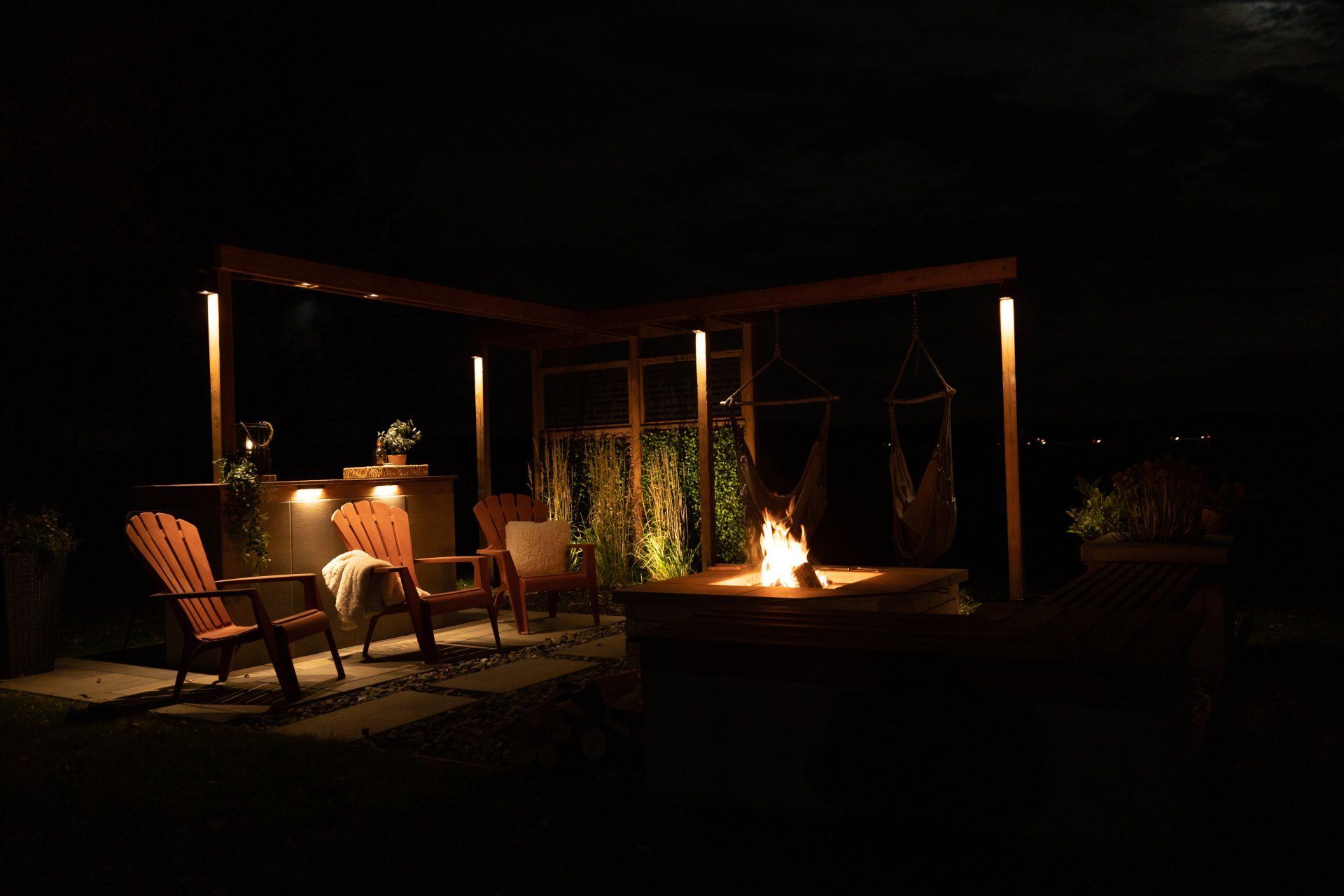 Une terrasse extérieure illuminée la nuit, avec des chaises en bois, un foyer et une pergola ornée de guirlandes lumineuses.