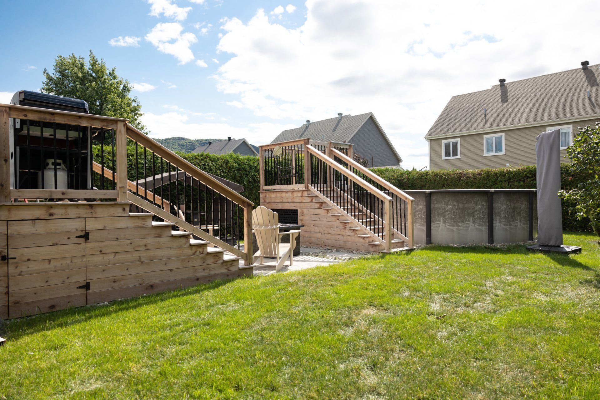 Par une journée ensoleillée, le jardin arrière comprend une terrasse en bois avec des escaliers menant à une piscine, avec des maisons en arrière-plan.