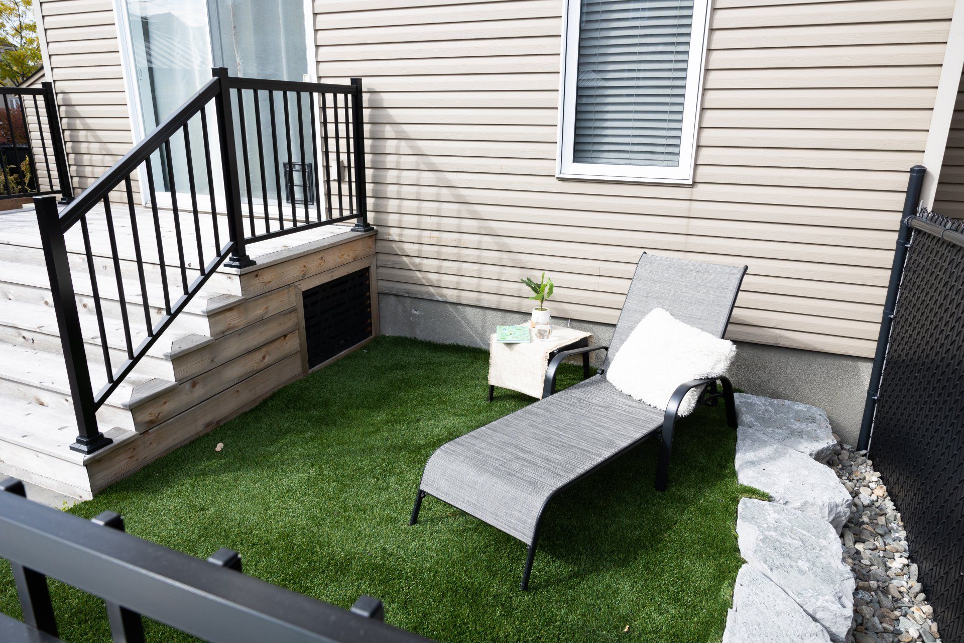 Un fauteuil gris avec un coussin blanc est installé sur une parcelle de gazon artificiel à côté d'une terrasse et d'une maison au bardage beige.