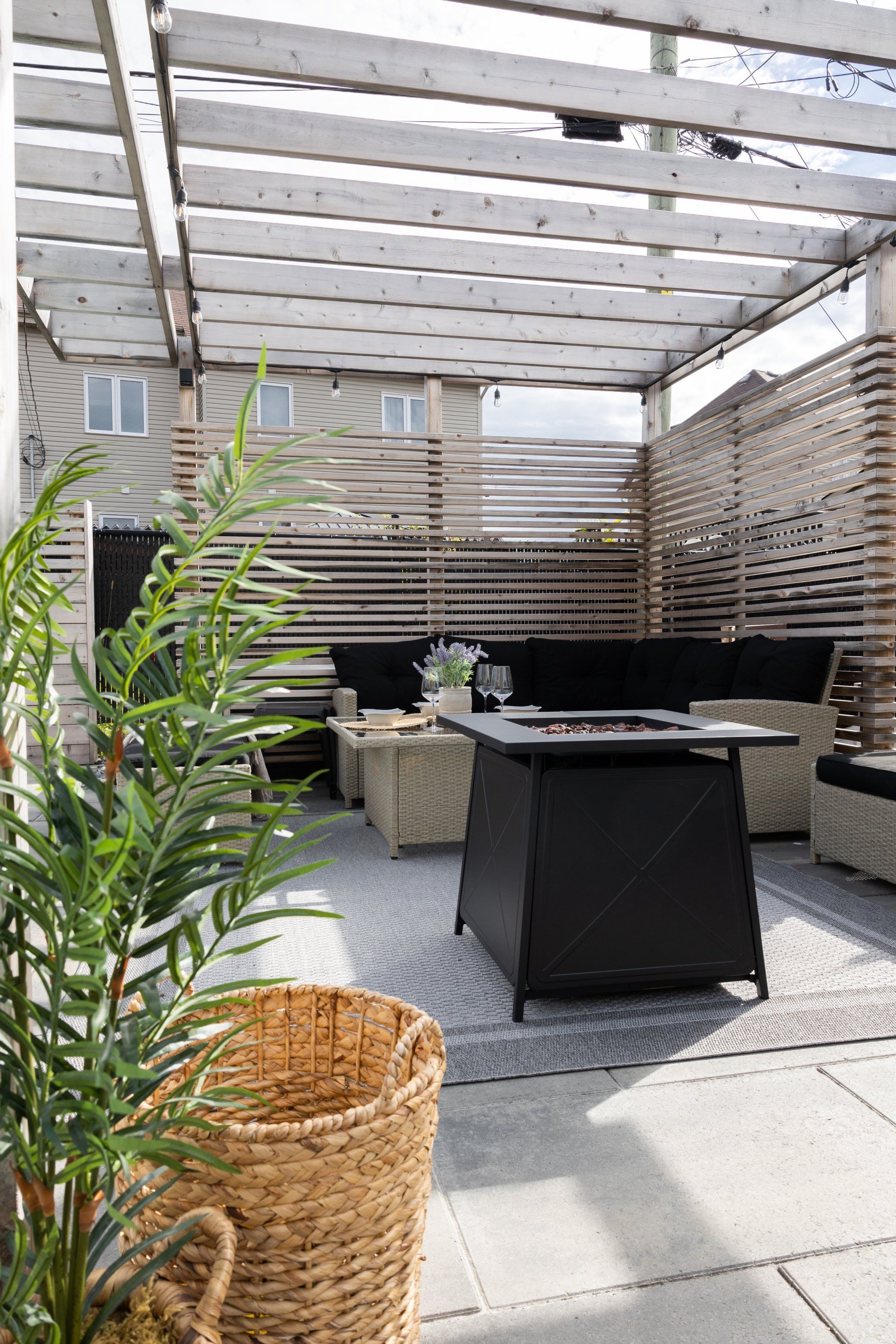 Terrasse extérieure avec pergola, mobilier de jardin en rotin clair, foyer extérieur foncé et panier tressé.