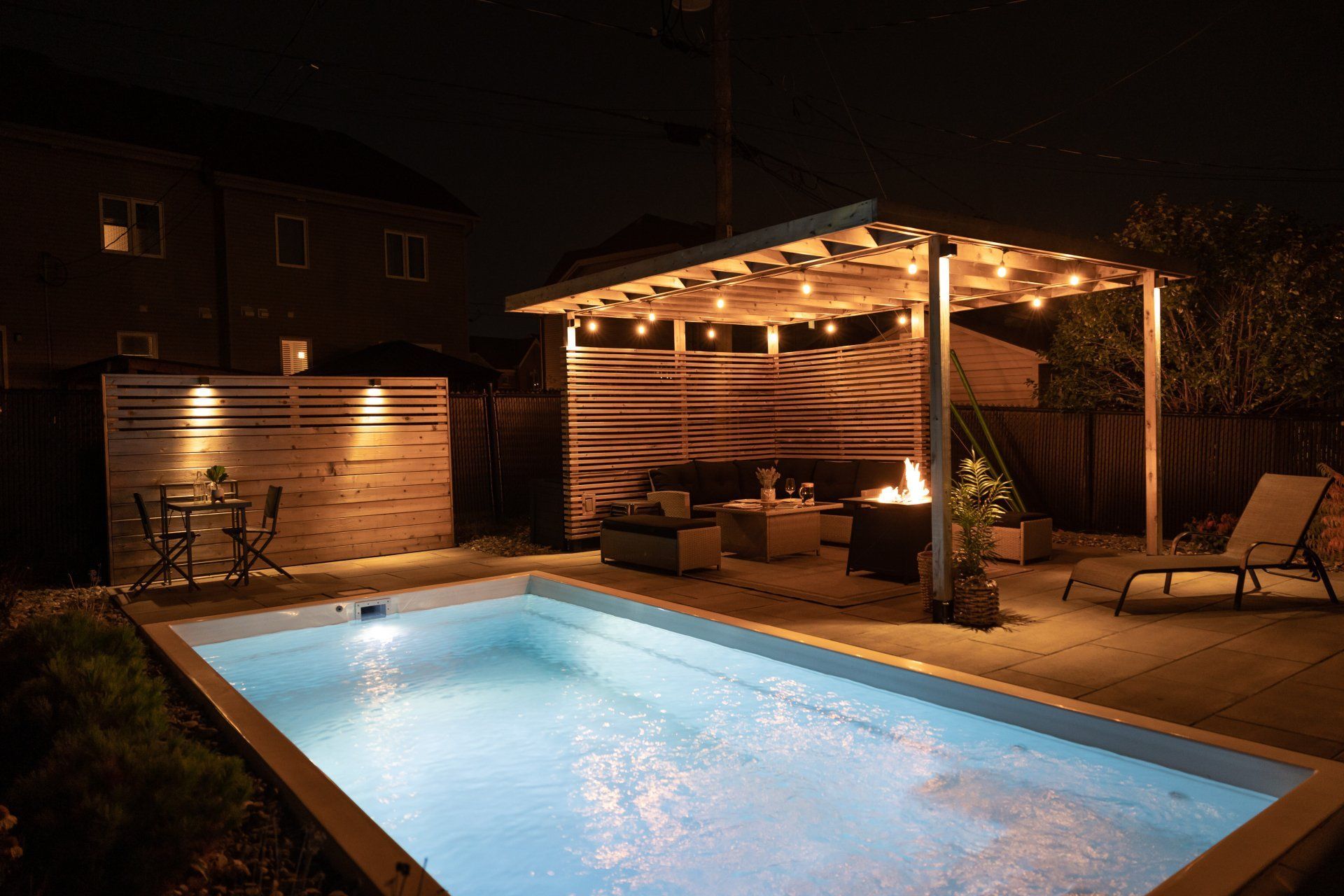 Une cour arrière illuminée la nuit, avec une piscine scintillante, un foyer, des meubles de patio et des guirlandes lumineuses sous une pergola.