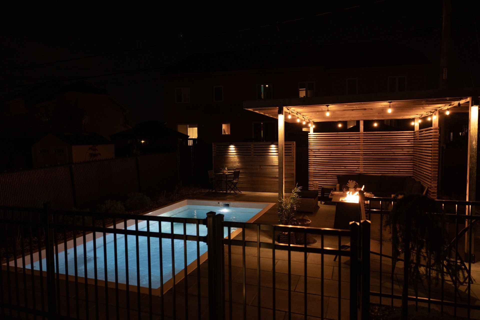 La nuit, dans un jardin sombre, une piscine brille d'un bleu éclatant, à côté d'une pergola illuminée par des guirlandes lumineuses et d'un foyer.