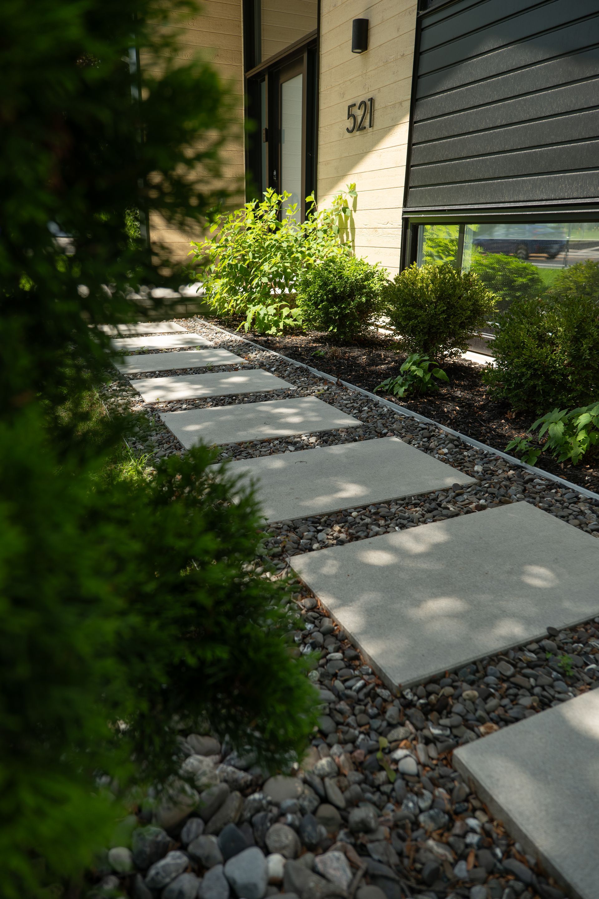 Un chemin de dalles rectangulaires en béton, entouré de pierres décoratives sombres, mène à l'entrée d'une maison moderne.
