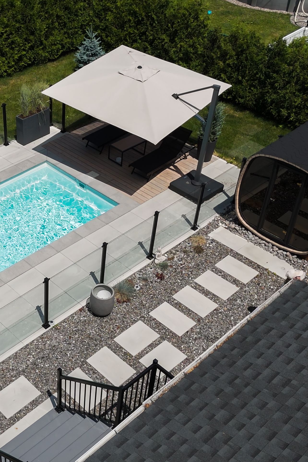 Vue plongeante sur un jardin moderne avec piscine, parasol carré, dalles de pierre et sauna.