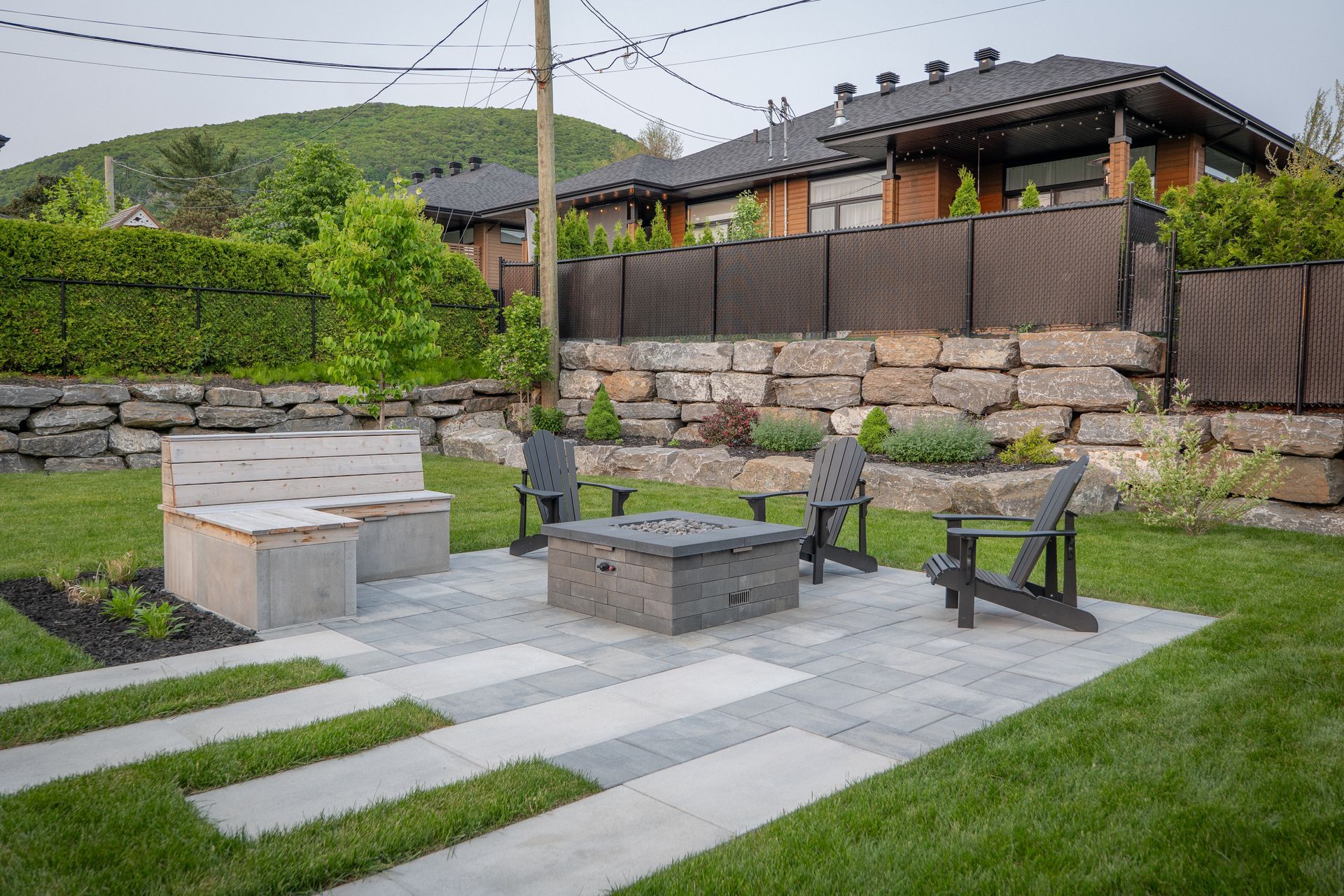 Une terrasse avec un foyer, des chaises Adirondack et un banc en pierre dans une cour arrière, agrémentée d'un mur de soutènement en pierre et de gazon.