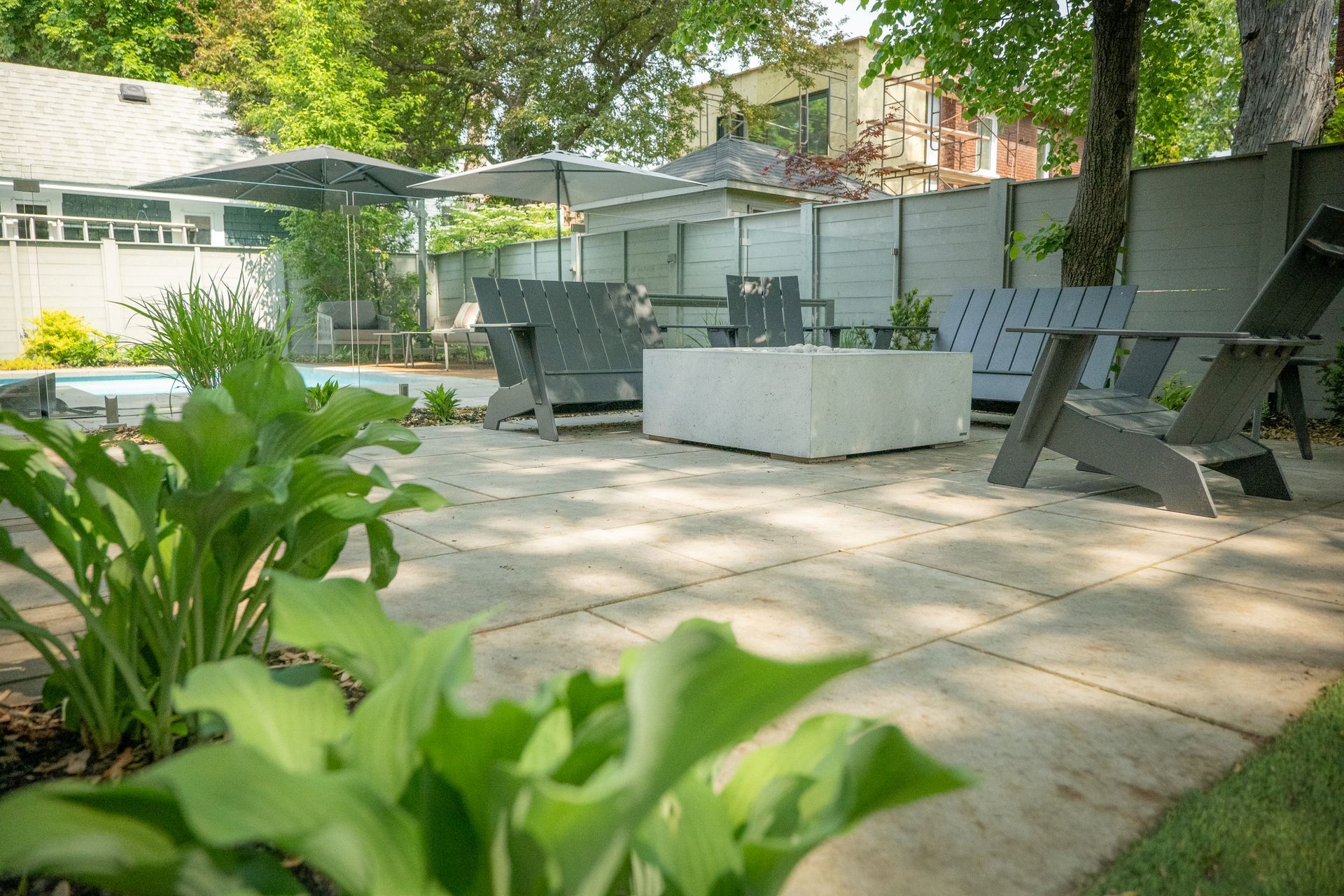 Une terrasse est agrémentée d'un foyer carré en béton, de chaises Adirondack grises, de parasols et d'une végétation luxuriante au premier plan.