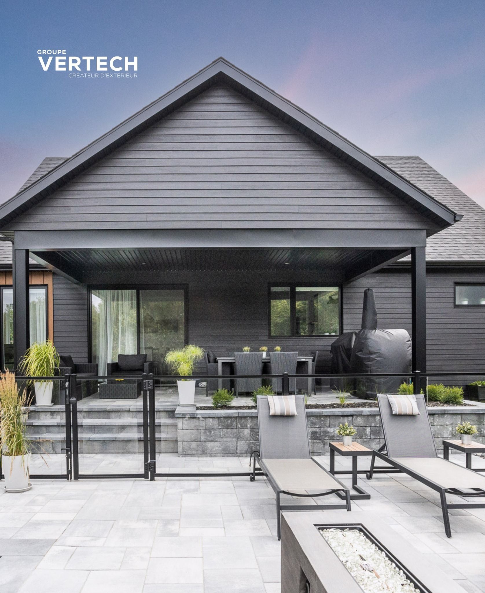 Une maison moderne gris foncé avec une terrasse couverte, un coin repas extérieur, des chaises longues et un foyer en pierre dans le jardin.