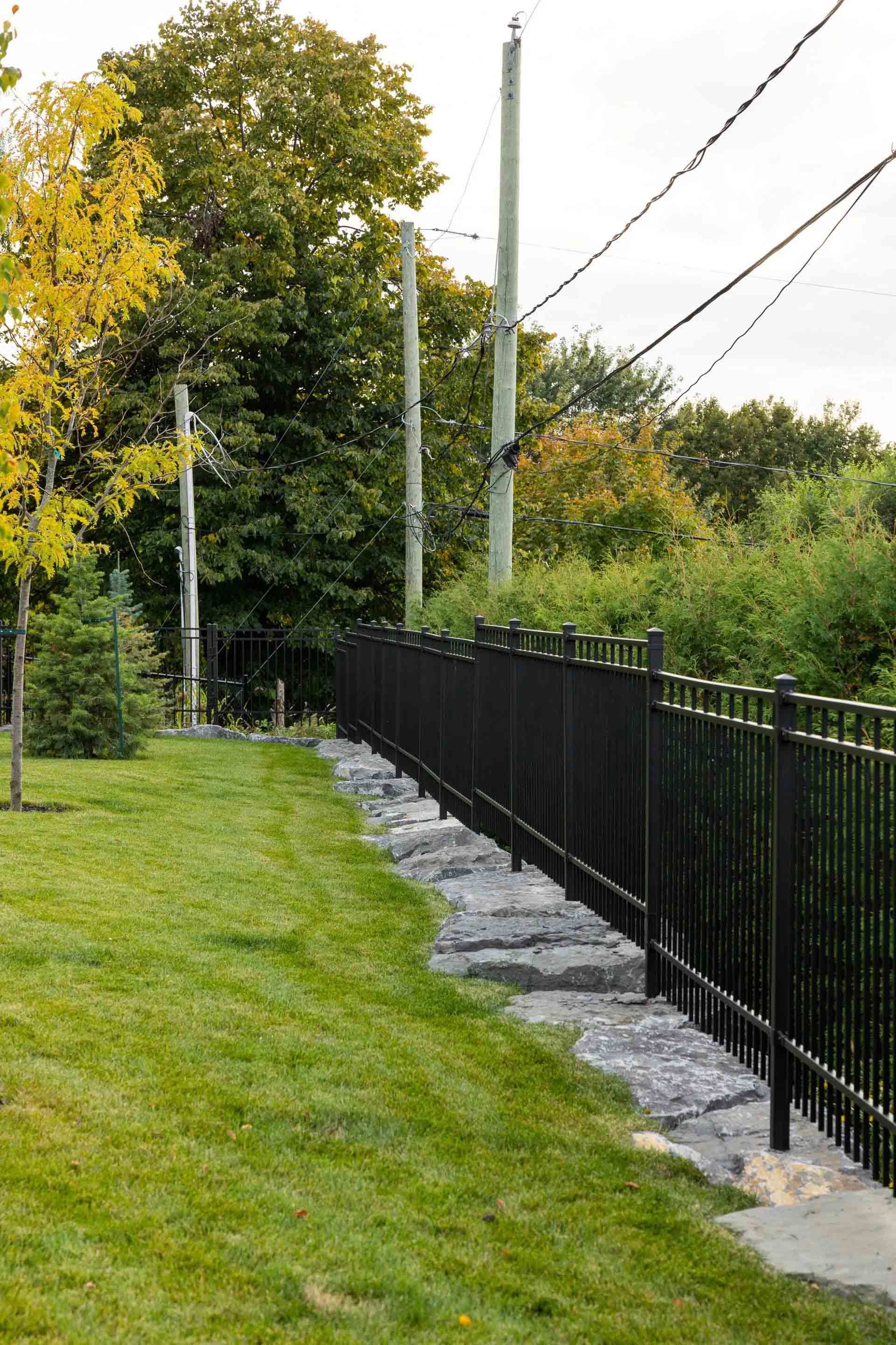 Une clôture en métal noir longe un mur de soutènement en pierre qui borde une pelouse verte, avec des poteaux électriques et des arbres à l'arrière.