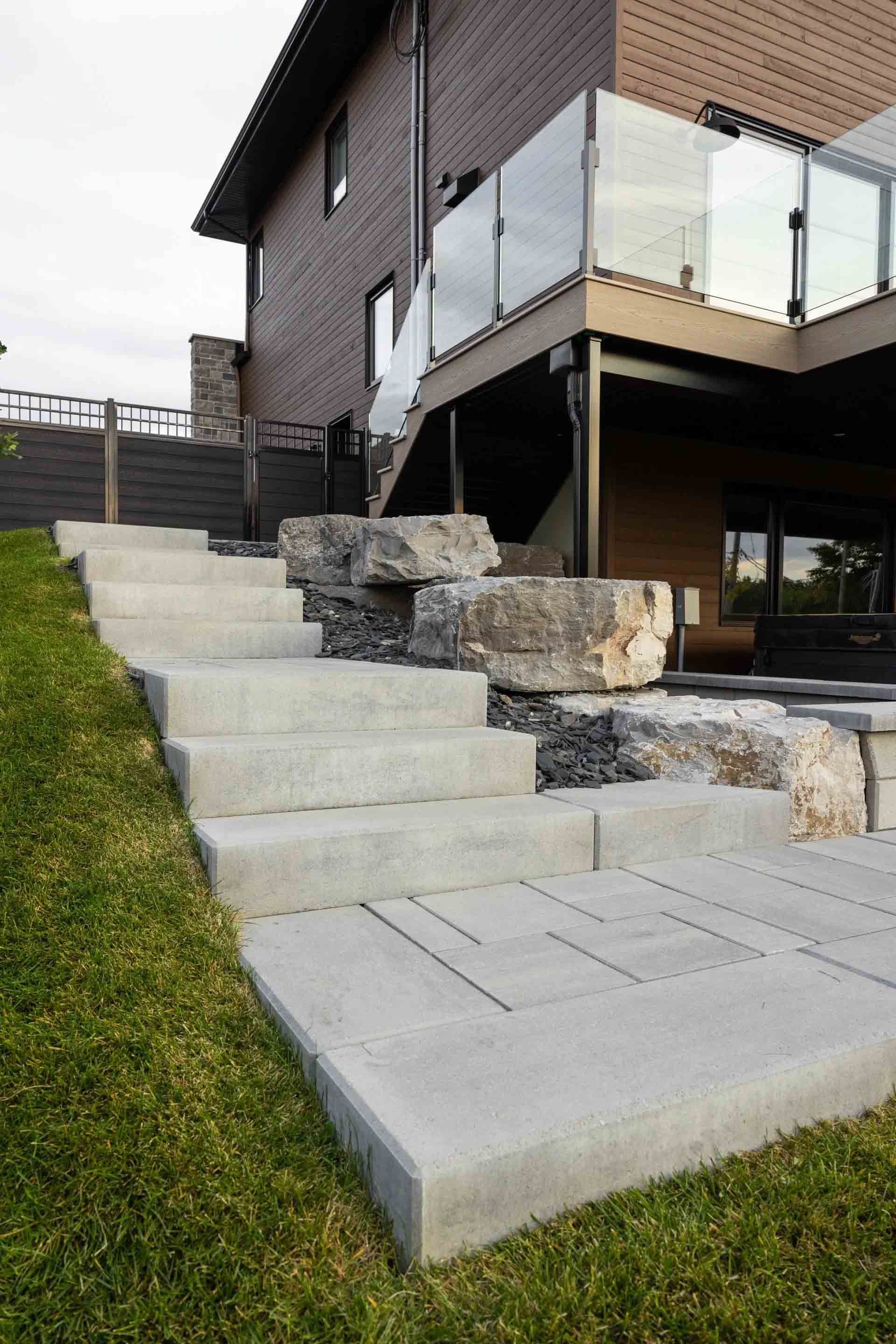 Un escalier extérieur en béton mène à une terrasse en bois avec une balustrade en verre, bordée de grosses pierres décoratives.