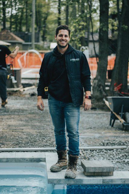 Une personne souriante, vêtue d'un gilet sombre et d'un jean, se tient près d'un chantier de construction avec une piscine.