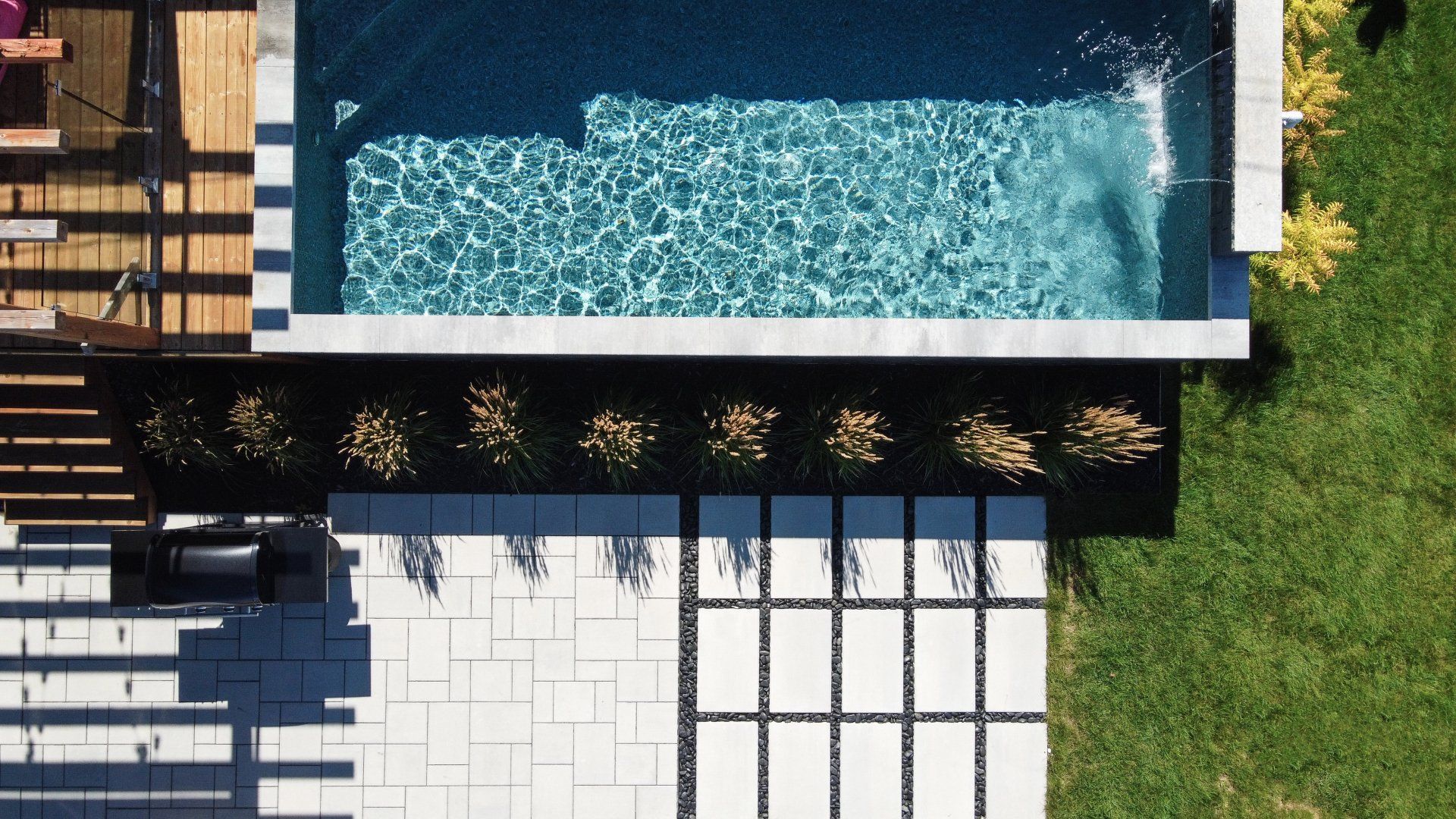 Vue aérienne d'une piscine moderne jouxtant une terrasse en pierre, une terrasse en bois, des parterres de fleurs et une pelouse.