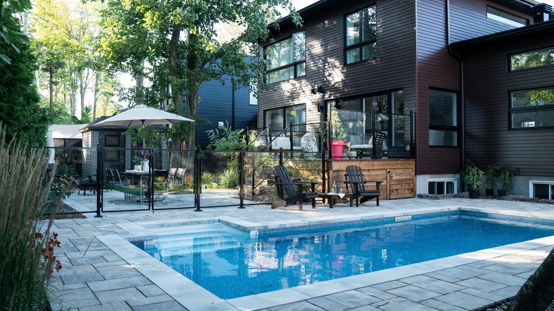 Une piscine rectangulaire entourée d'une terrasse en pierre, avec une maison moderne en bois sombre et des arbres en arrière-plan.