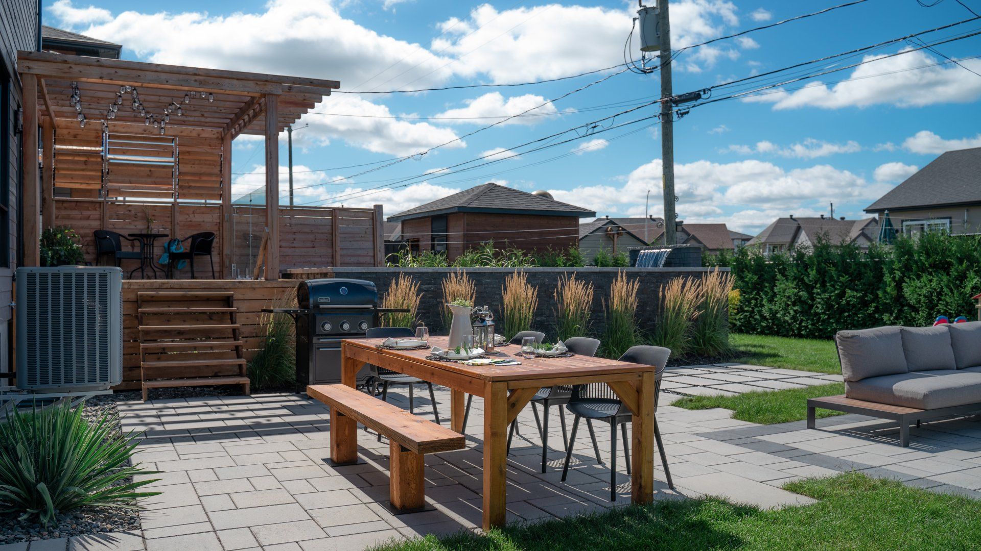 Une terrasse en bois avec une pergola et des escaliers, comprenant un ensemble de salle à manger, un barbecue et des meubles de salon sur une cour arrière pavée.