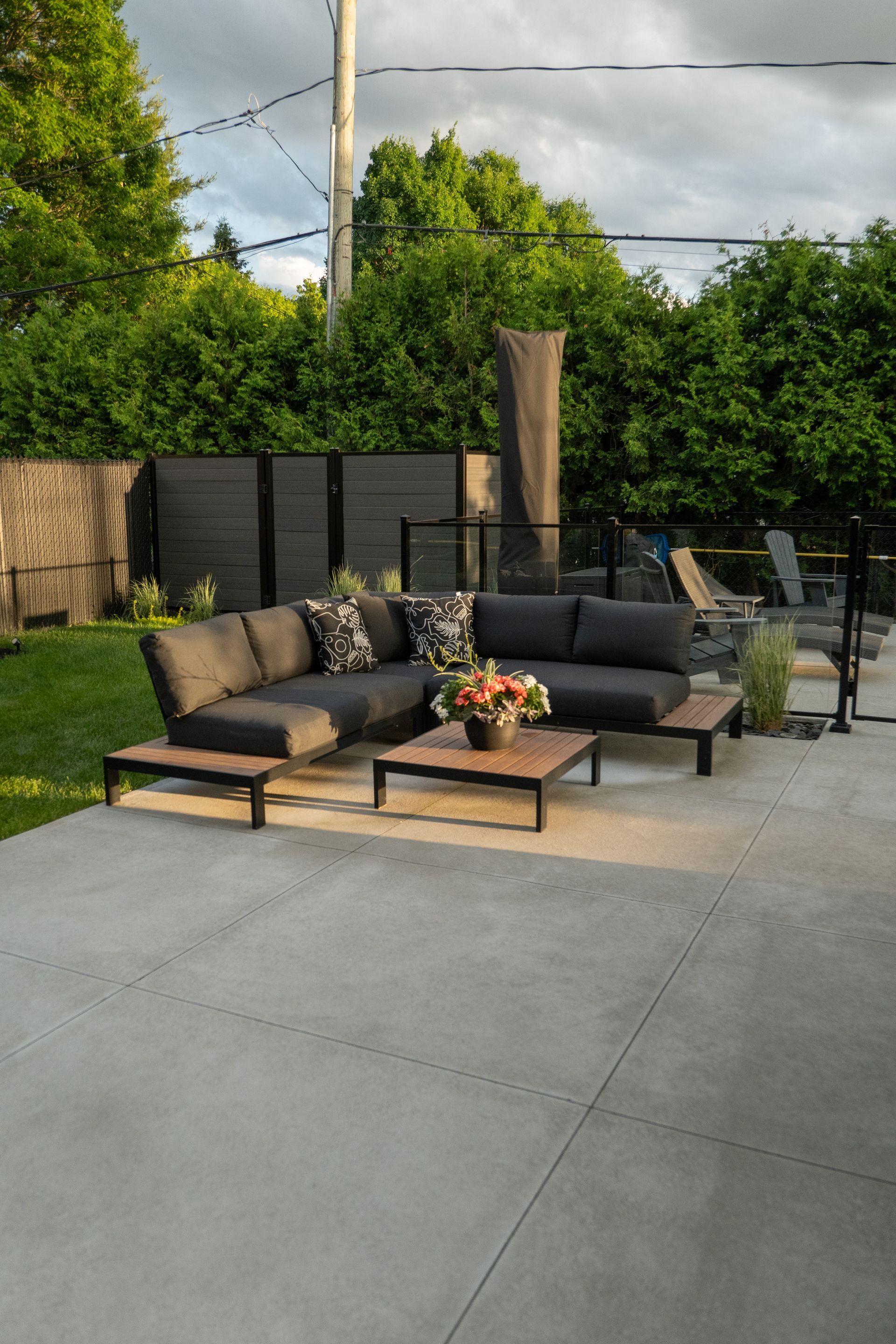 Un canapé d'angle gris en forme de L, agrémenté de coussins à motifs, et une table basse en bois sont installés sur une terrasse en béton, au cœur d'un jardin verdoyant.