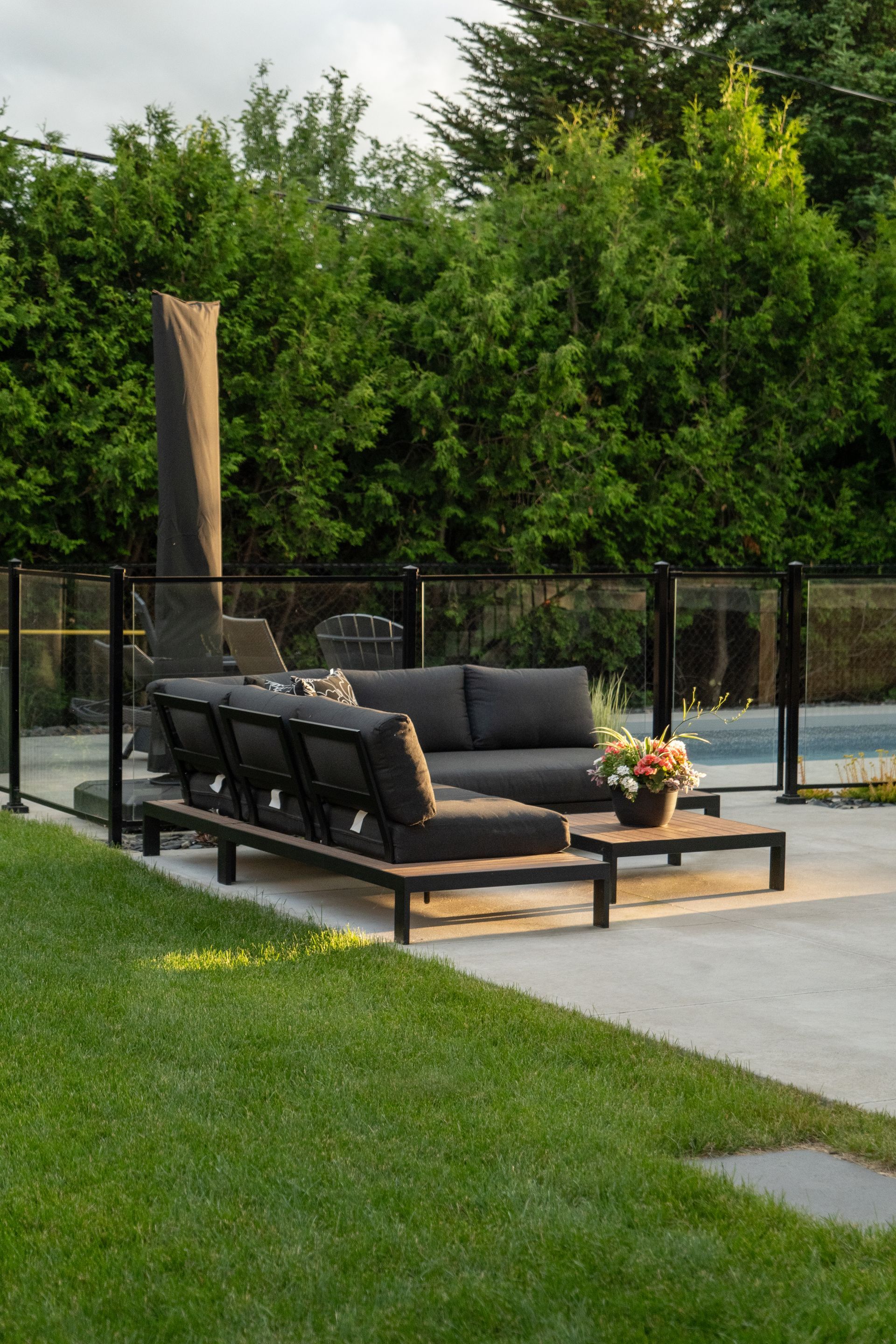 Un canapé d'angle noir d'extérieur avec des coussins foncés et une table basse assortie, sur une terrasse au bord d'une piscine entourée d'une clôture en verre.