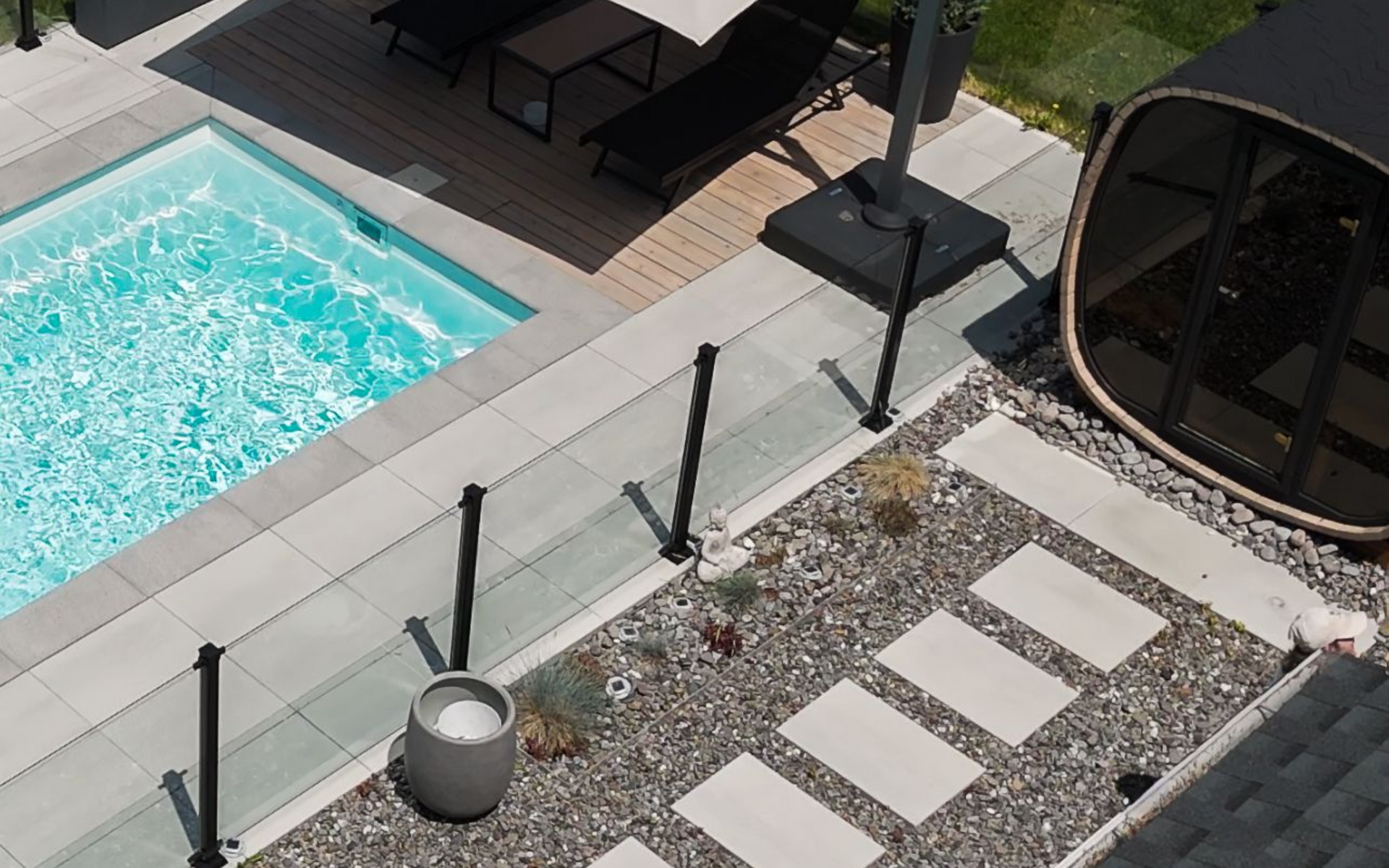 Une piscine rectangulaire est installée sur une terrasse en pierre avec un ensemble de salle à manger, des chaises longues et un pool house moderne, le tout adossé à une maison bleue.
