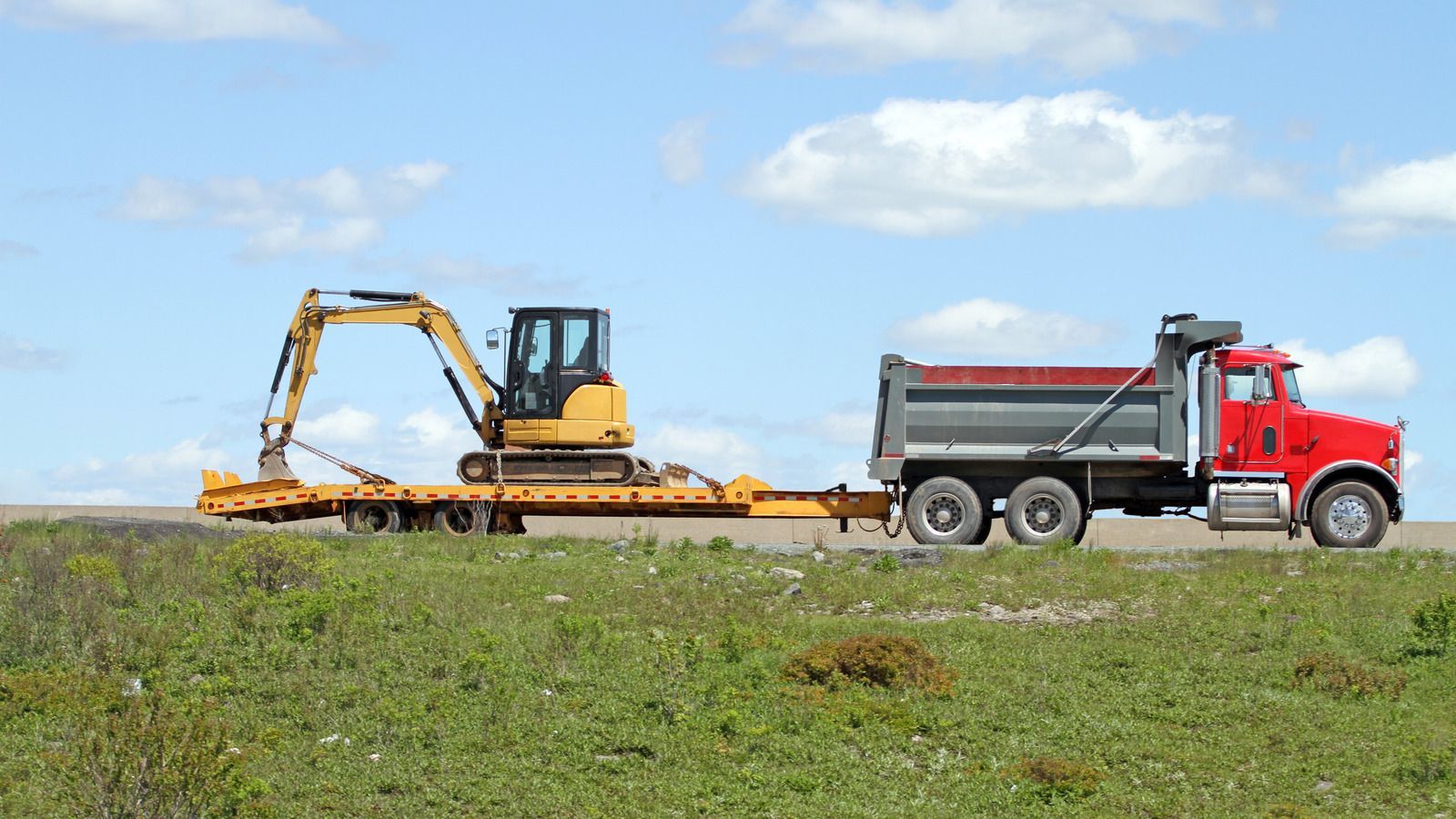 Red dump truck towing a flatbed trailer with a yellow excavator on a road, against a blue sky.