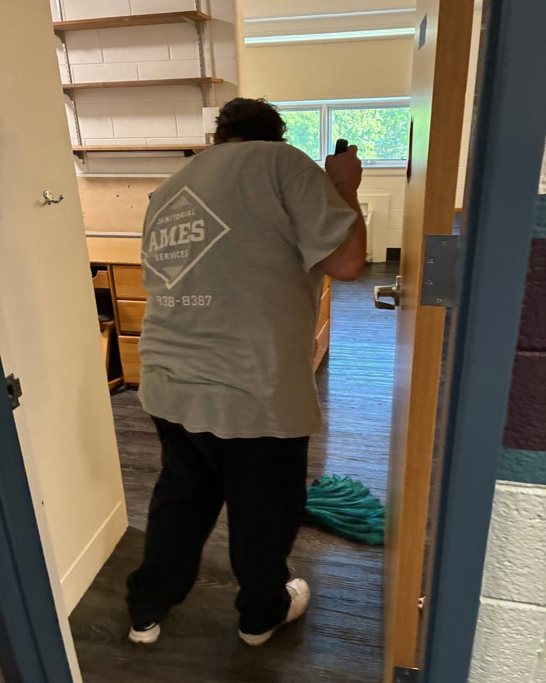 Person vacuuming a carpeted room, viewed from a doorway, with shelves and a window in the background.