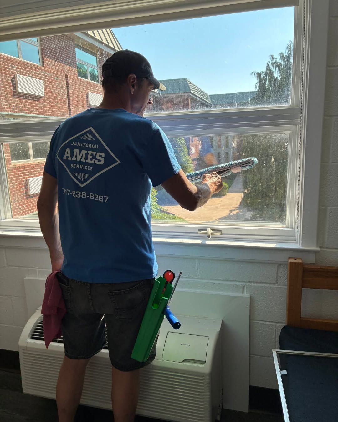 Worker in blue shirt caulking a window inside a room with a brick building outside.