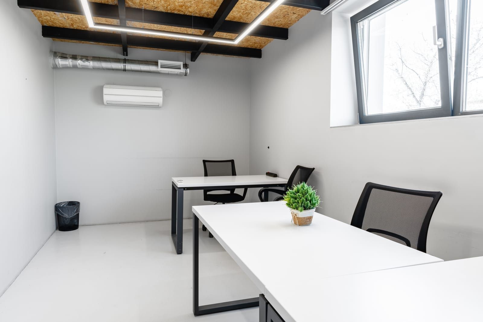Minimal office with white desks, black chairs, exposed ceiling, wall AC, and a small window.