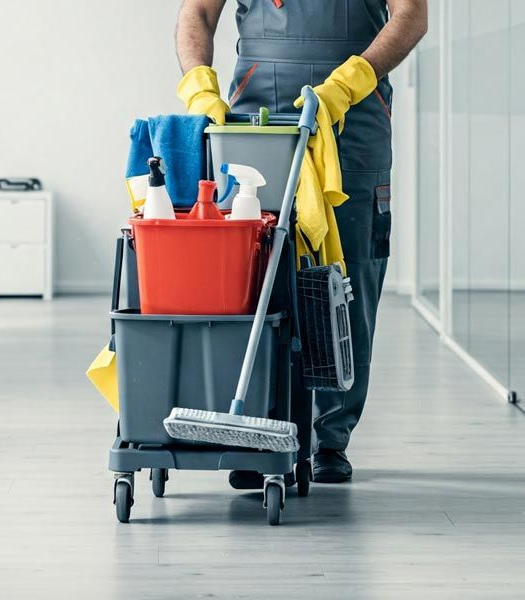 A cleaner in work uniform pushes a wheeled cart filled with cleaning supplies, including buckets, spray bottles, and a mop.