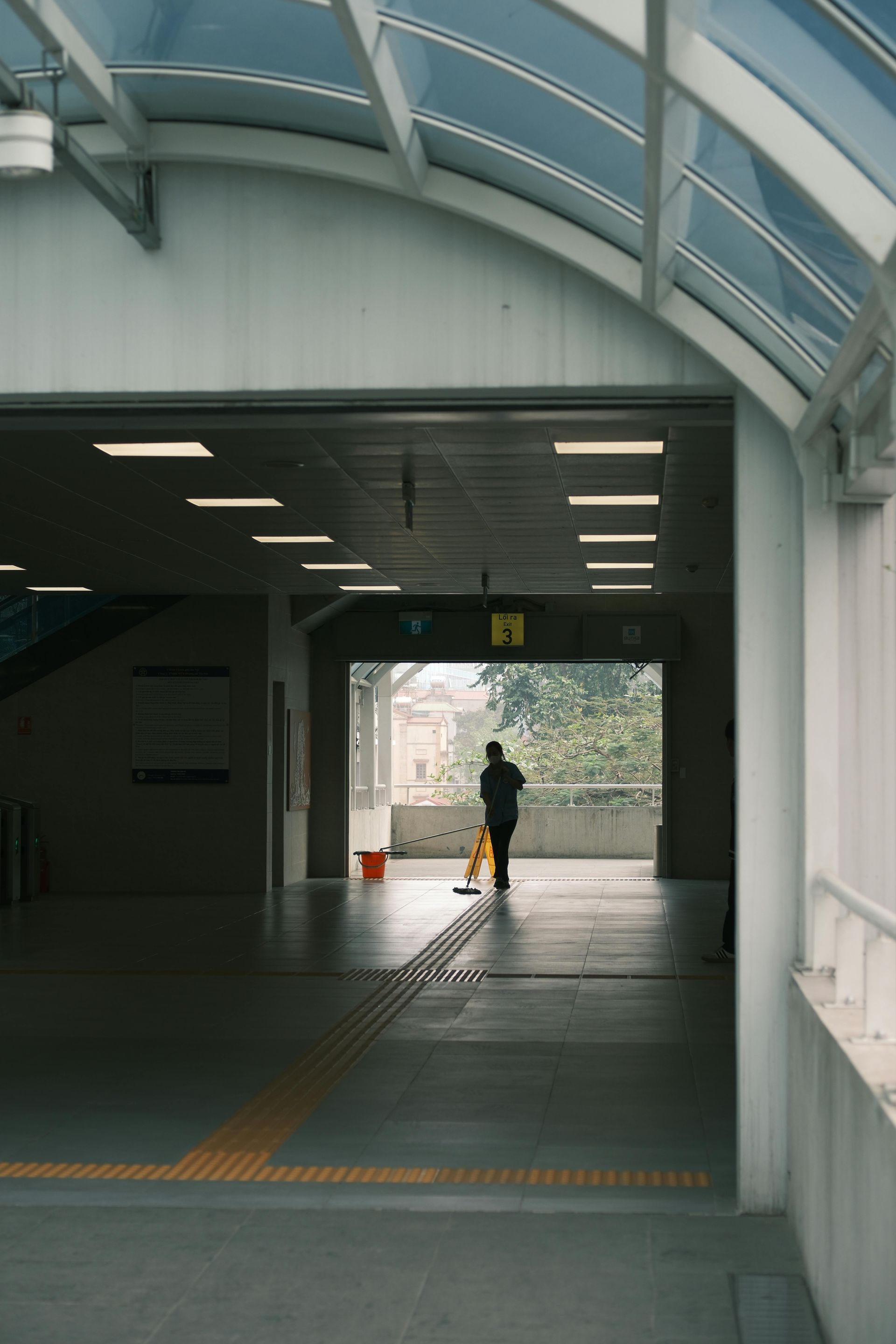 A person cleans the floor of a brightly lit, arched indoor pedestrian walkway with a yellow caution sign nearby.