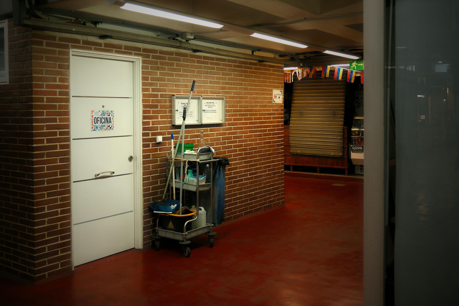 A cleaning cart sits in a hallway with a white door against a brick wall, adjacent to an open space with red flooring.