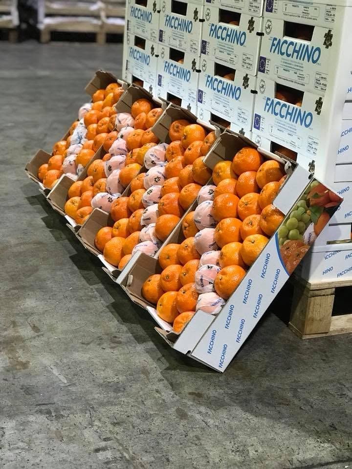 Scatole di arance impilate in un magazzino, con cassette per la frutta bianche e arancioni sullo sfondo.