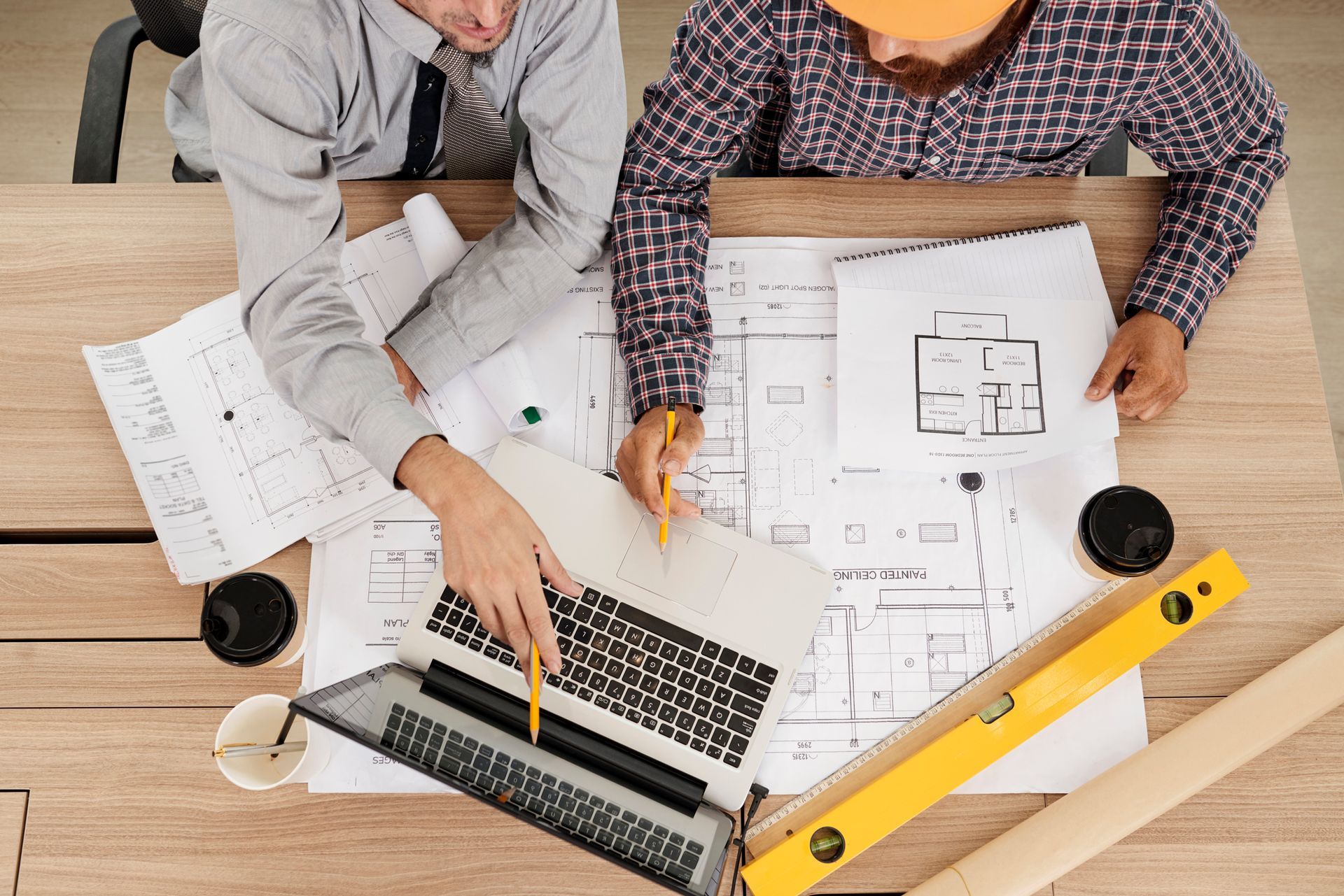 Two men are sitting at a table looking at blueprints and a laptop.