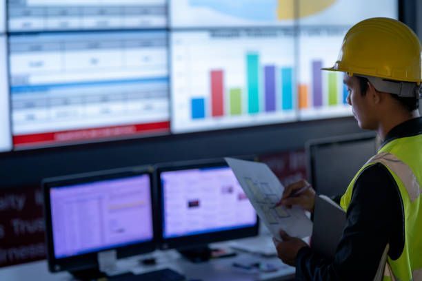 A man in a hard hat is looking at a computer screen while holding a piece of paper.