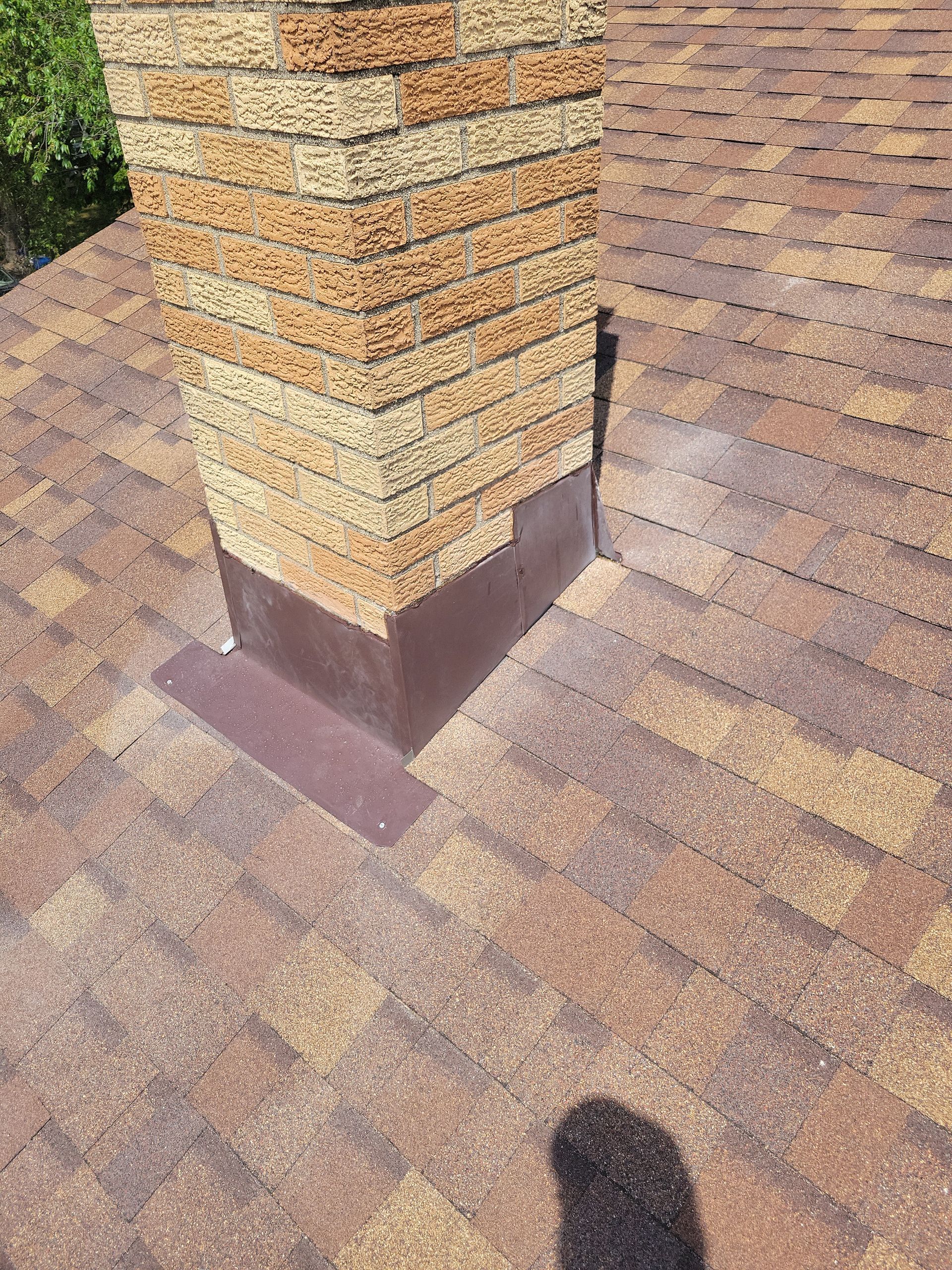 A brick chimney on a shingled roof with dark brown metal flashing installed around its base.