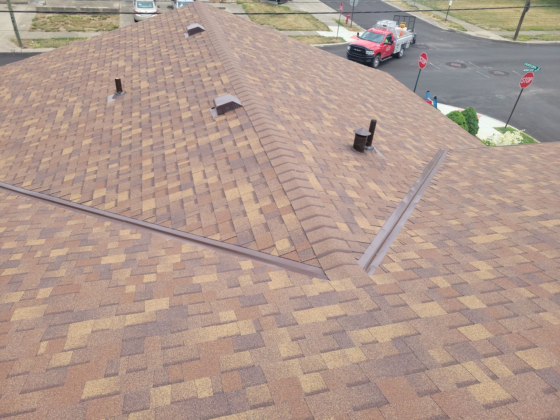 An elevated view of a brown shingled roof with metal valleys, vents, and a street with a vehicle and stop signs below.
